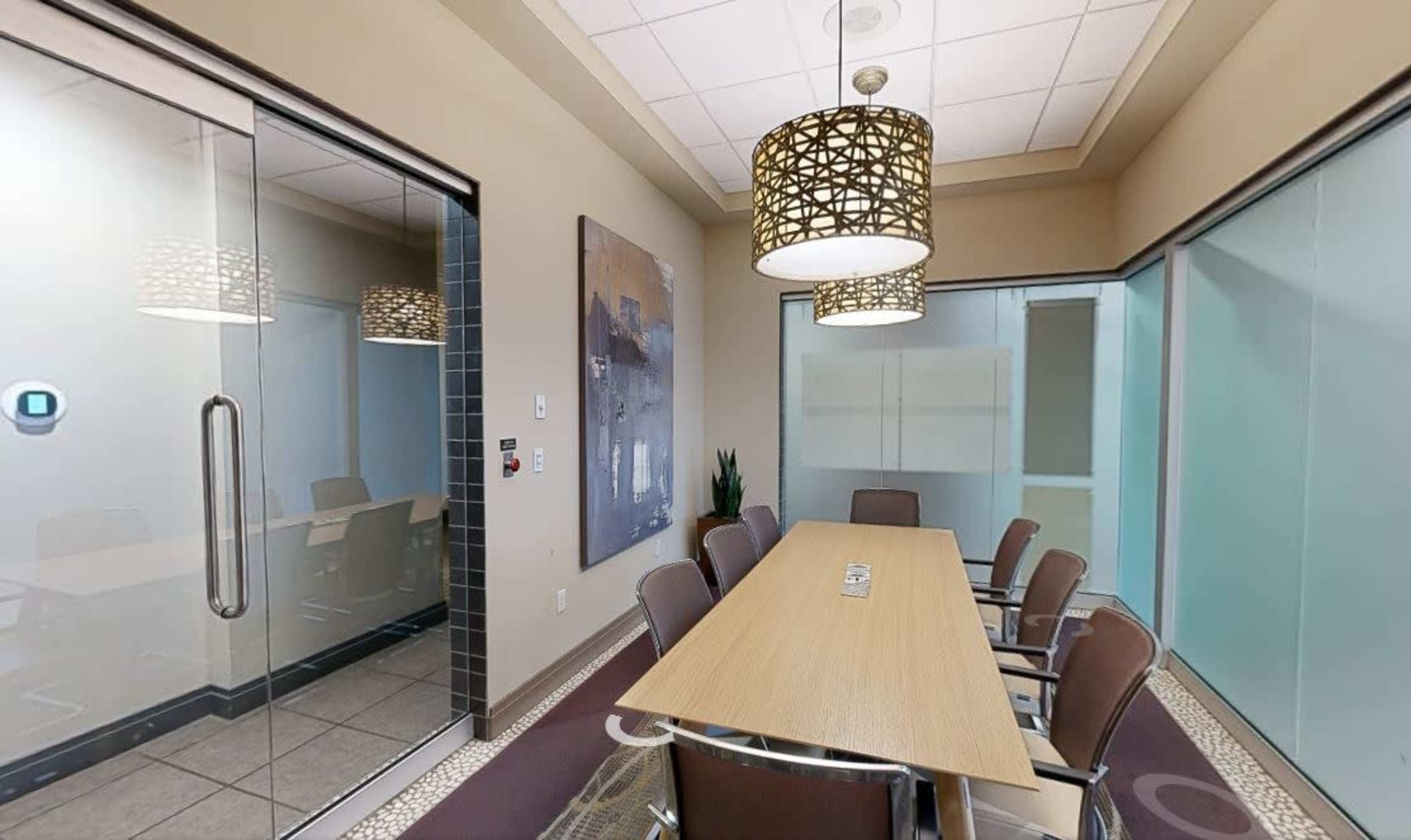 A modern conference room features a long wooden table surrounded by chairs, with decorative light fixtures and glass partitions.