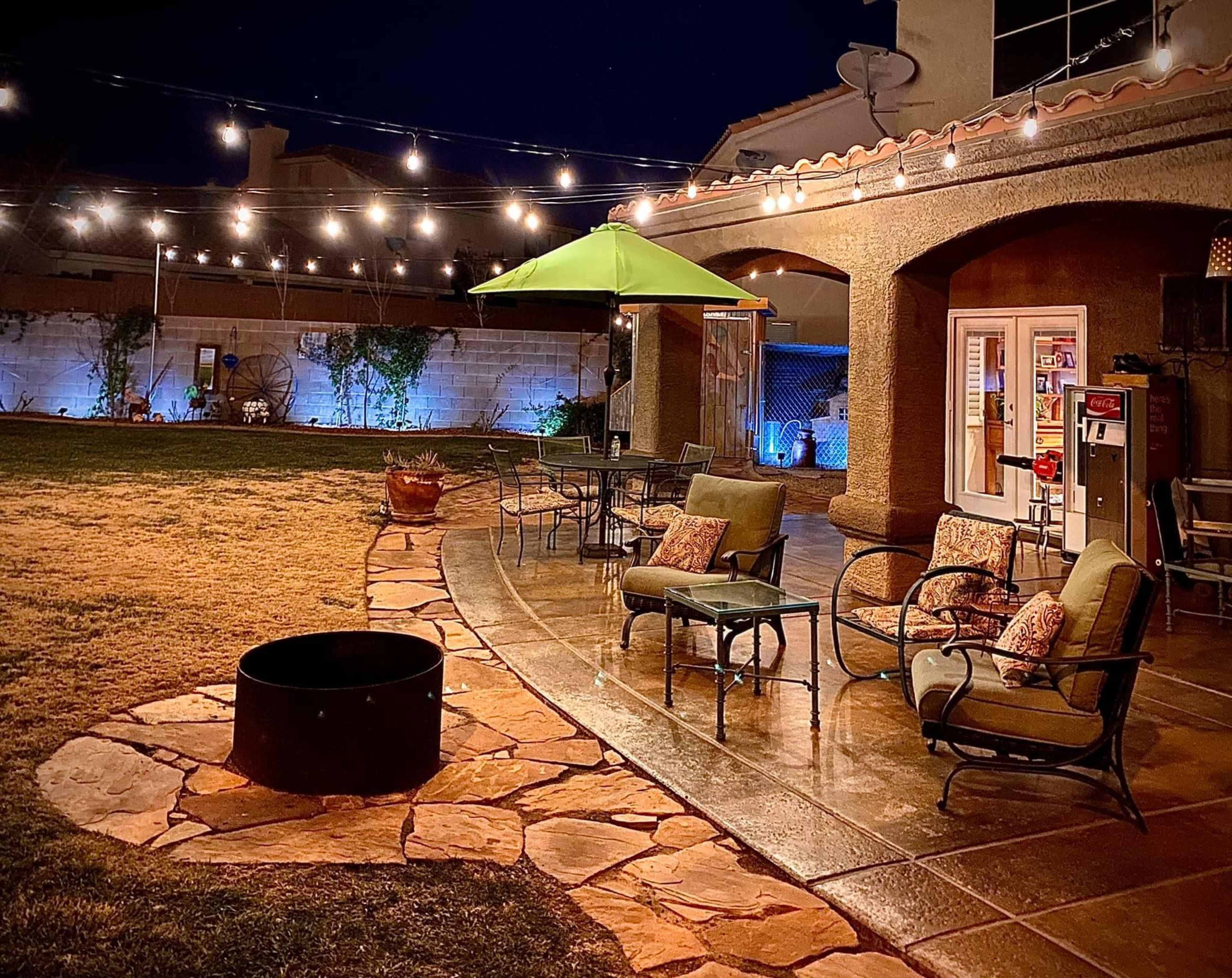 A backyard patio at night with string lights overhead, a green umbrella, seating arrangements, and a stone fire pit.