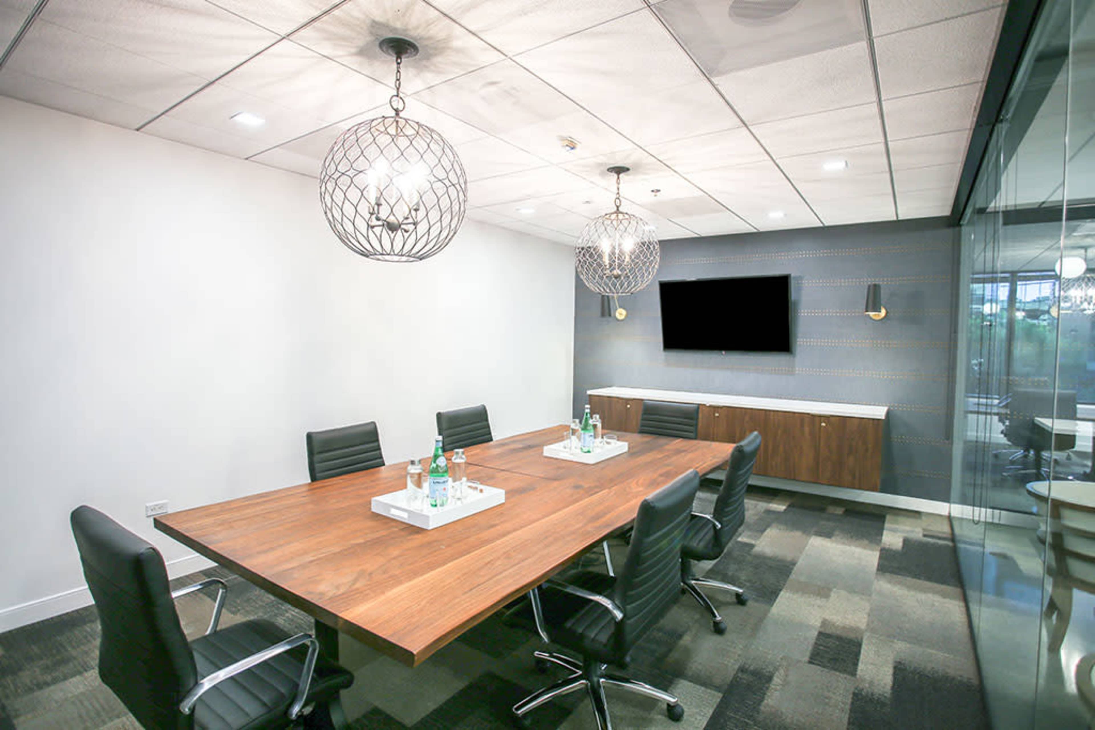 A modern conference room features a large wooden table surrounded by black chairs, with decorative pendant lights and a television on the wall.