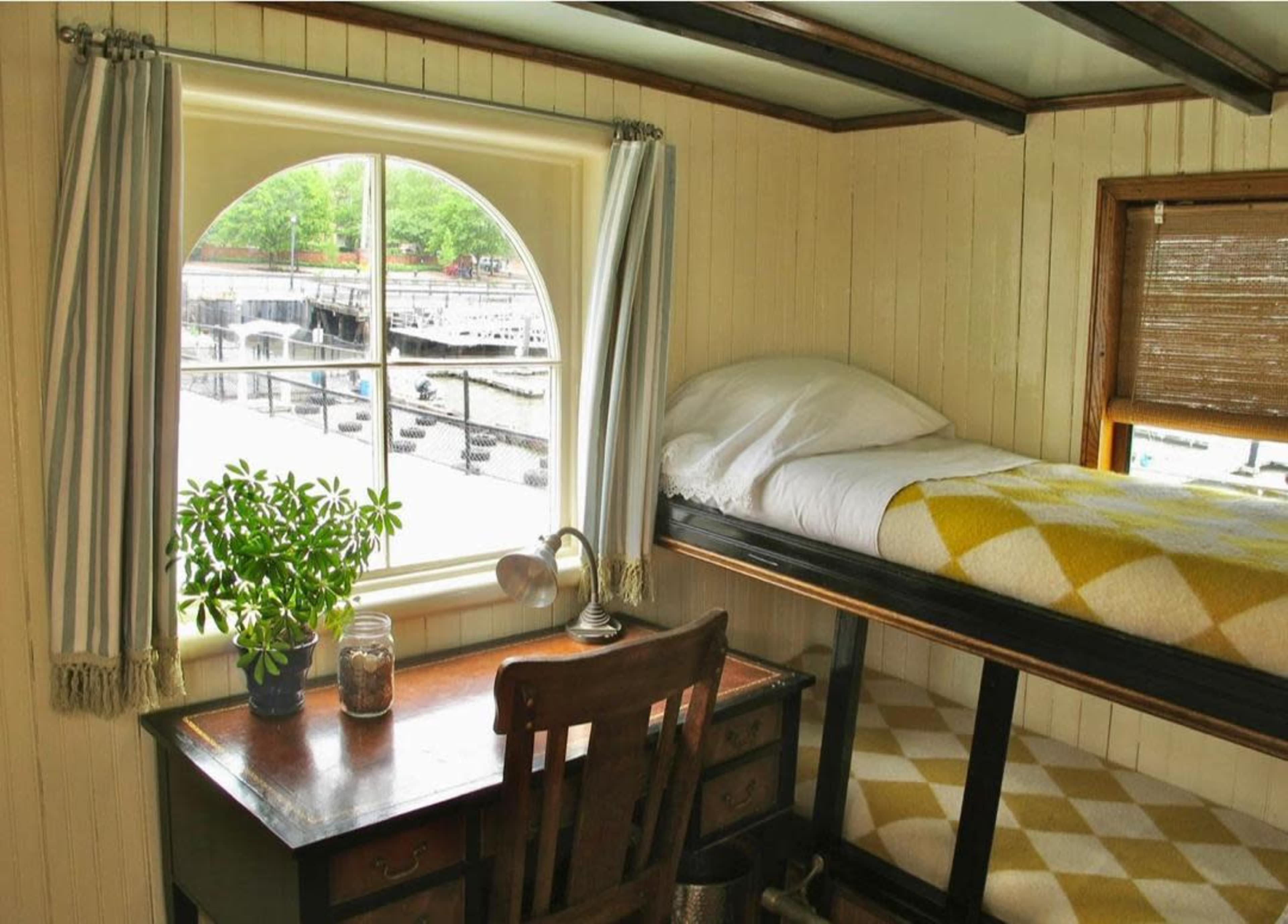 The interior of a small room features a bunk bed, a desk with a lamp, and a large arched window overlooking a waterfront scene.