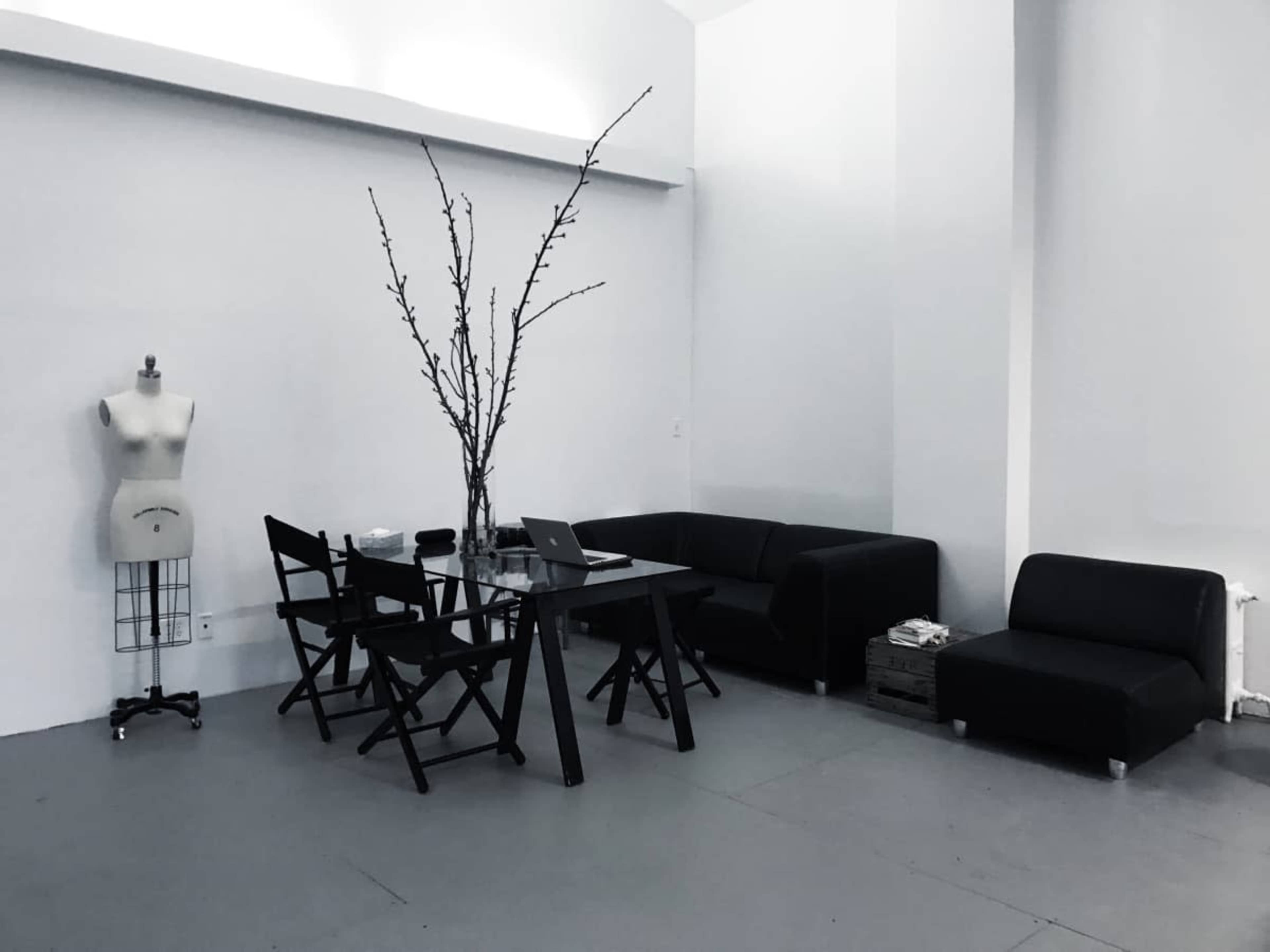 The image shows a minimalistic indoor space featuring a clear glass table surrounded by black chairs, a black sofa, and a mannequin, with tall branches arranged in a vase as a centerpiece.