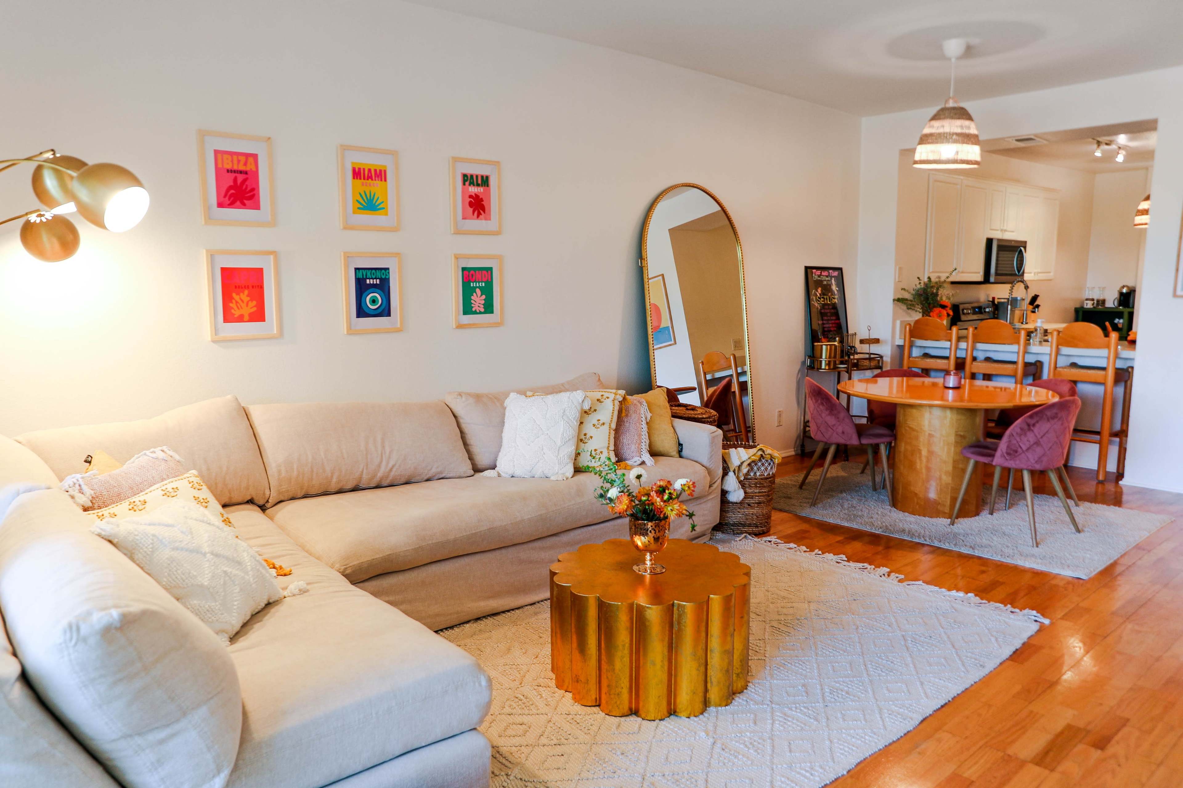 A bright living room with a beige sectional sofa, a round gold coffee table, colorful framed artwork on the wall, and a dining area with a round table and chairs.