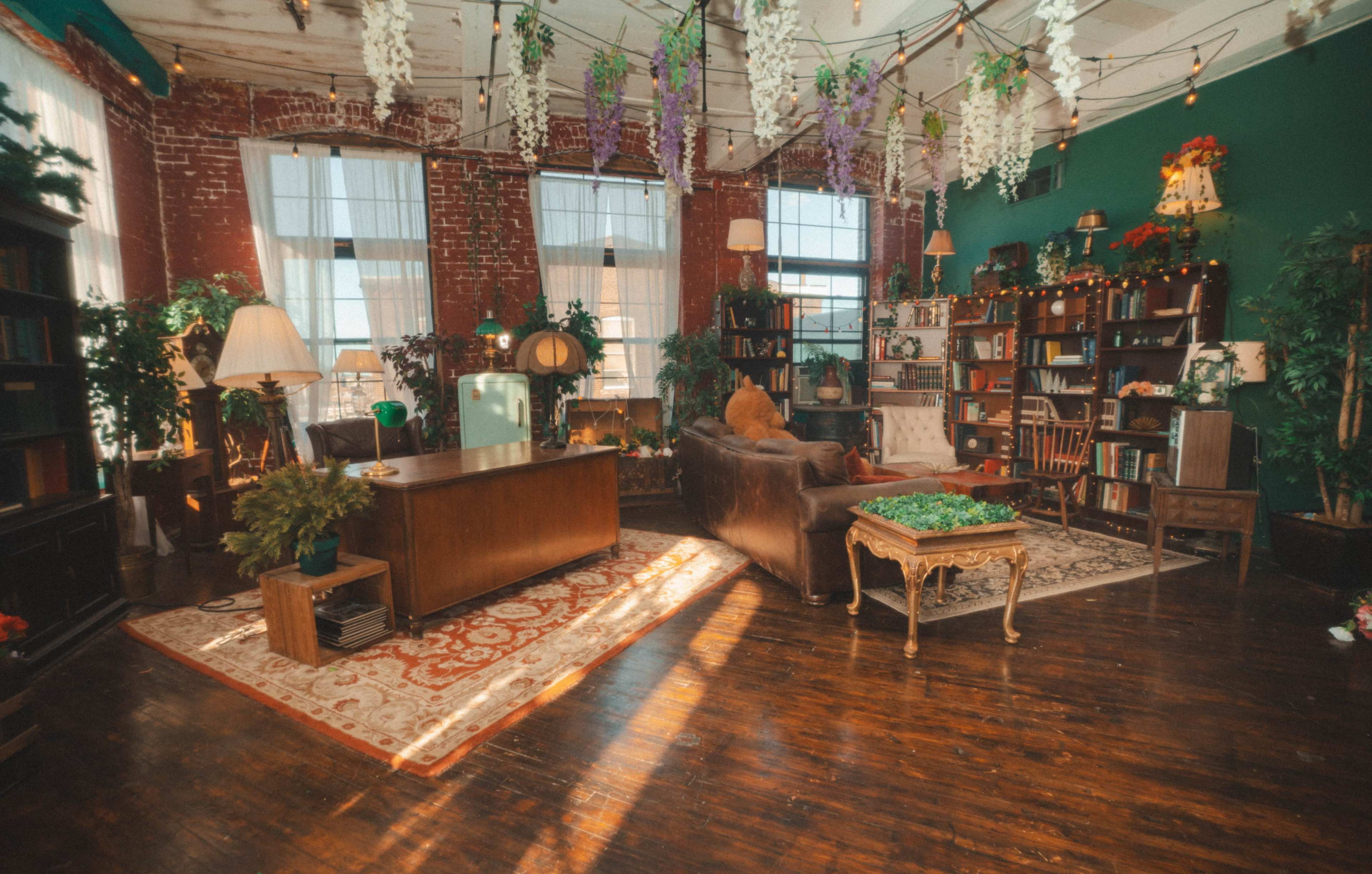 The image shows a bright, eclectic room featuring exposed brick walls, a mix of bookshelves filled with plants, and a comfortable seating area with a vintage desk and decor.