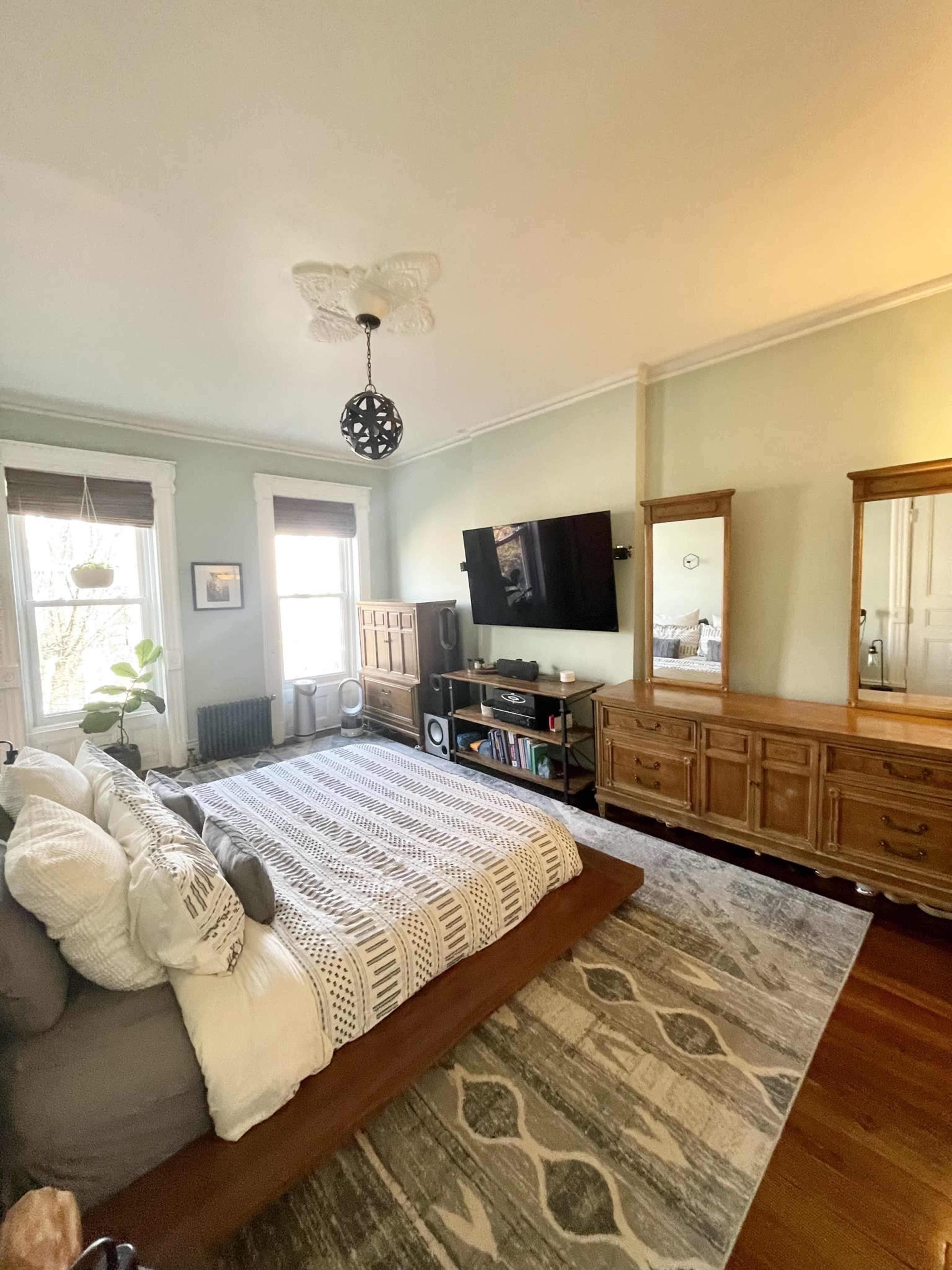A cozy bedroom with a large bed, two windows with shades, a wooden dresser and mirror set, and a television mounted on the wall.