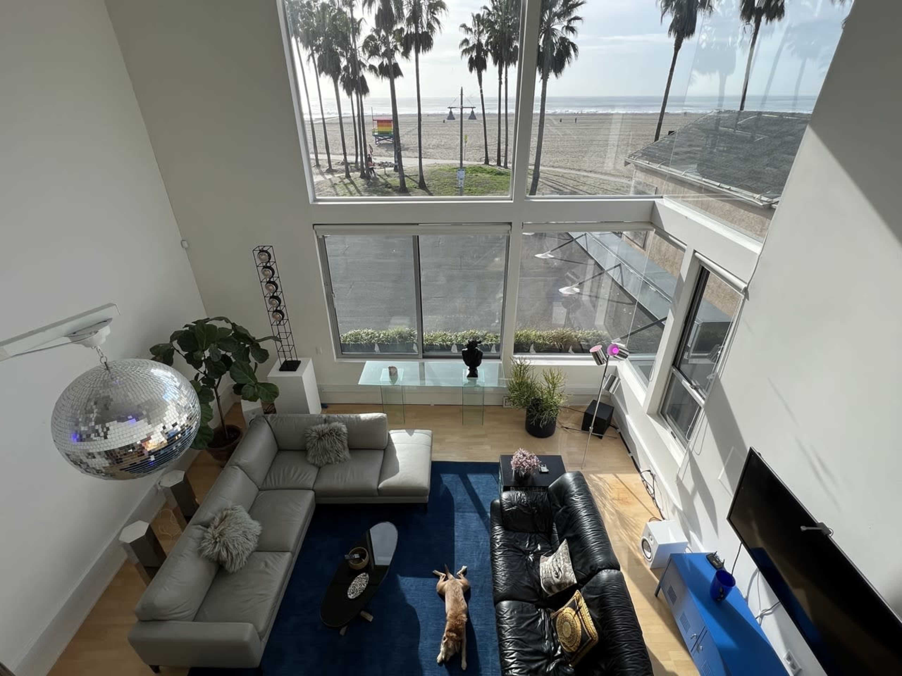 A spacious living room features a large sofa, a coffee table, and a view of the beach with palm trees through expansive windows.