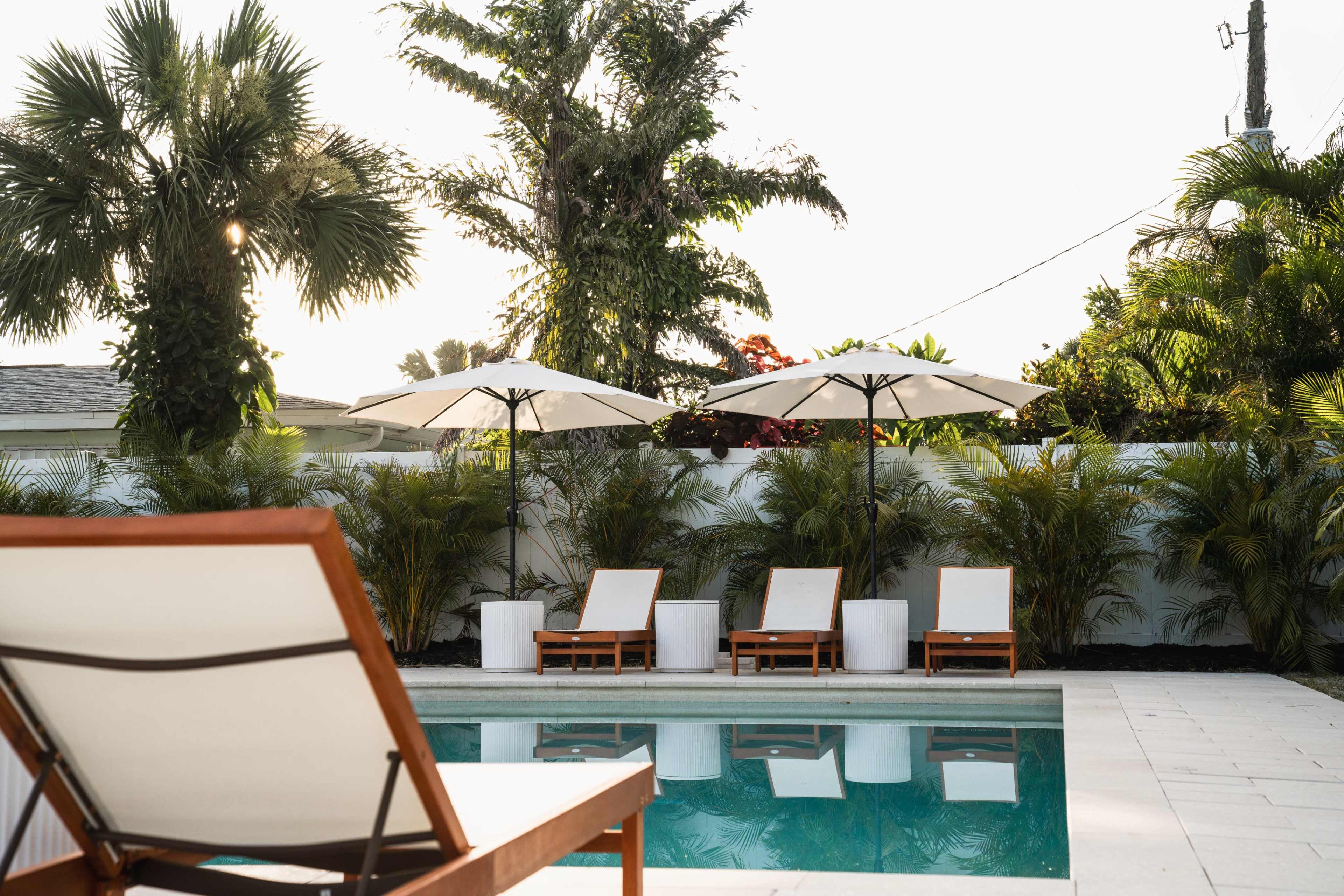 A lounge chair is positioned near a swimming pool surrounded by umbrellas and tropical plants.