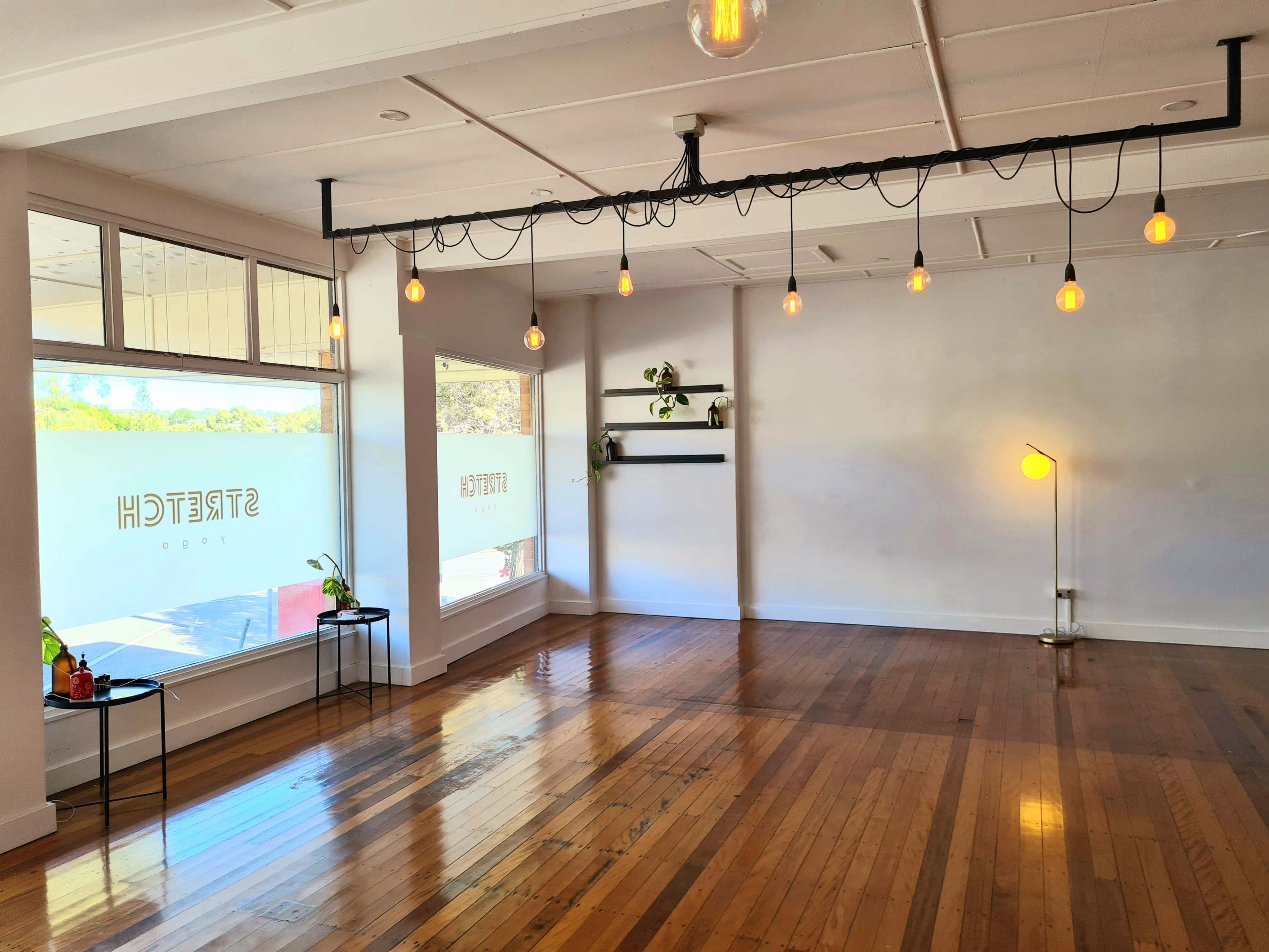 The image shows a spacious, empty room with wooden floors, large windows, and minimalist lighting fixtures hanging from the ceiling.