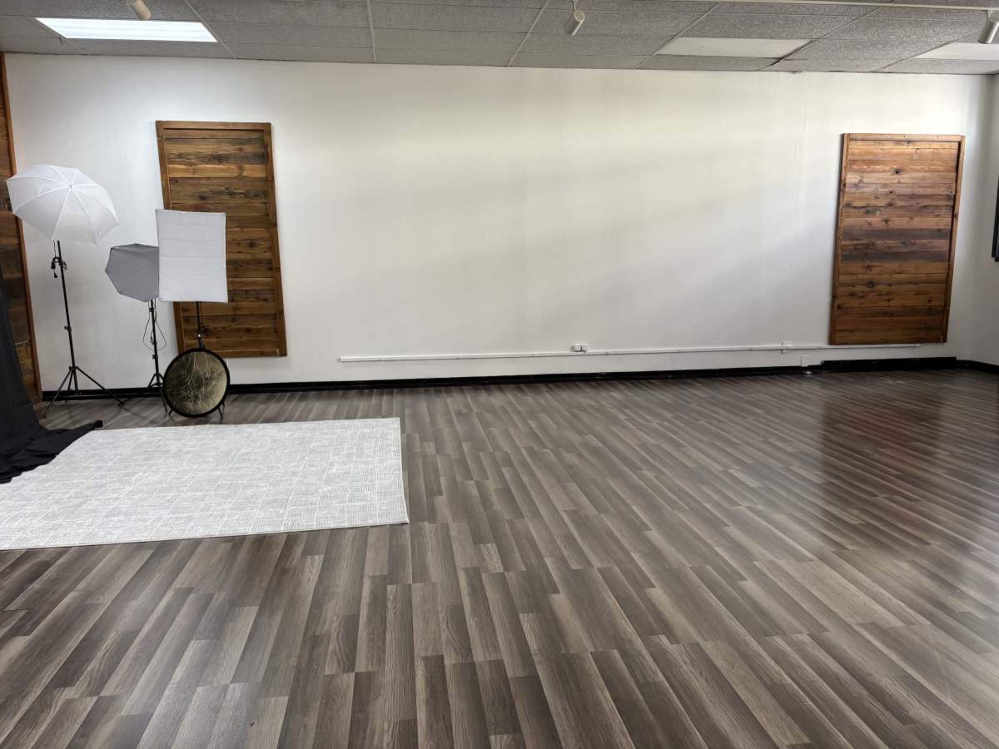 The image shows an empty room with wooden flooring, two large wooden wall panels, and photography lighting equipment set up in one corner.