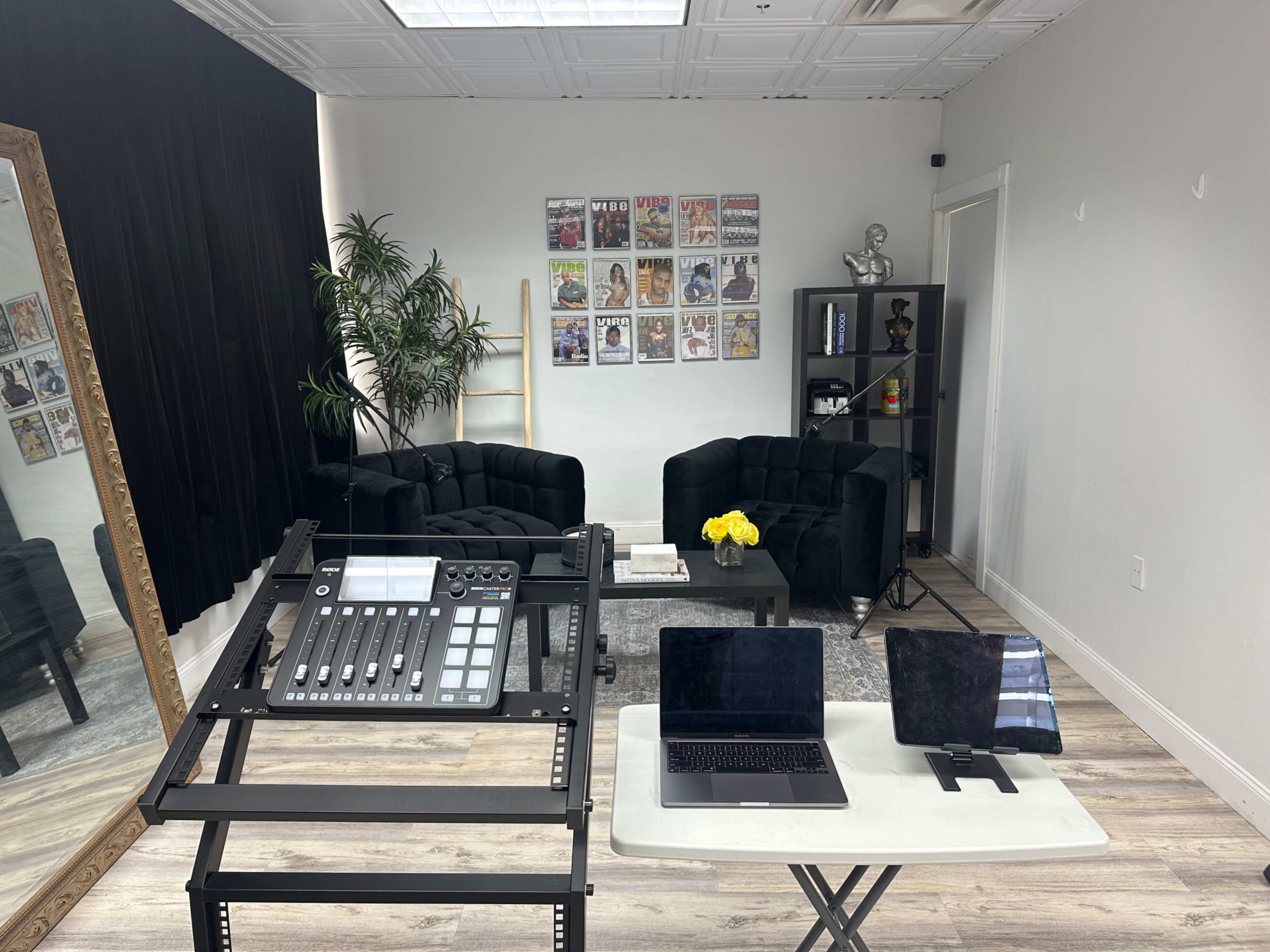 The image depicts a modern studio space featuring a black lounge area, a table with electronic equipment, and wall decorations of framed magazines.