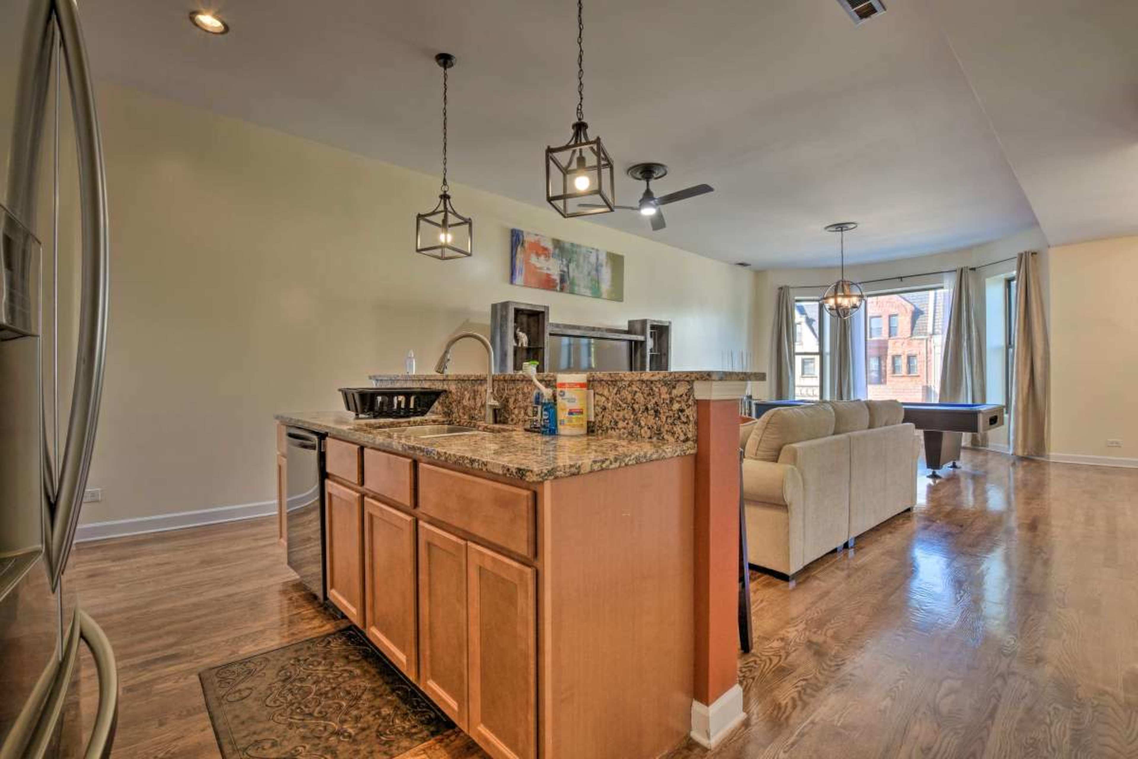 A kitchen area features wooden cabinets and a granite countertop, opening into a spacious living room with large windows.