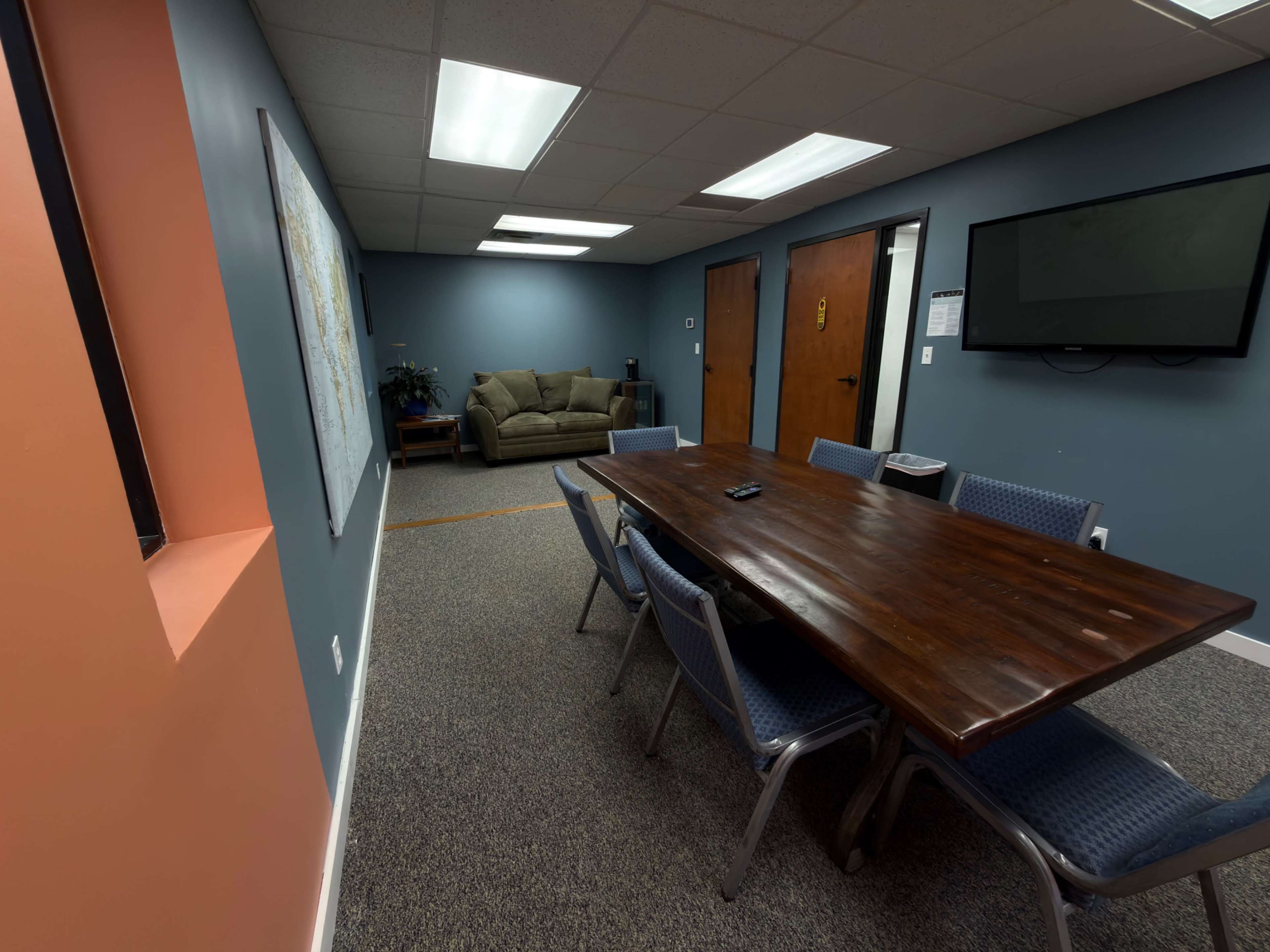 The image shows a conference room with a large wooden table surrounded by blue chairs, leading to a small lounge area with a couch in the background.