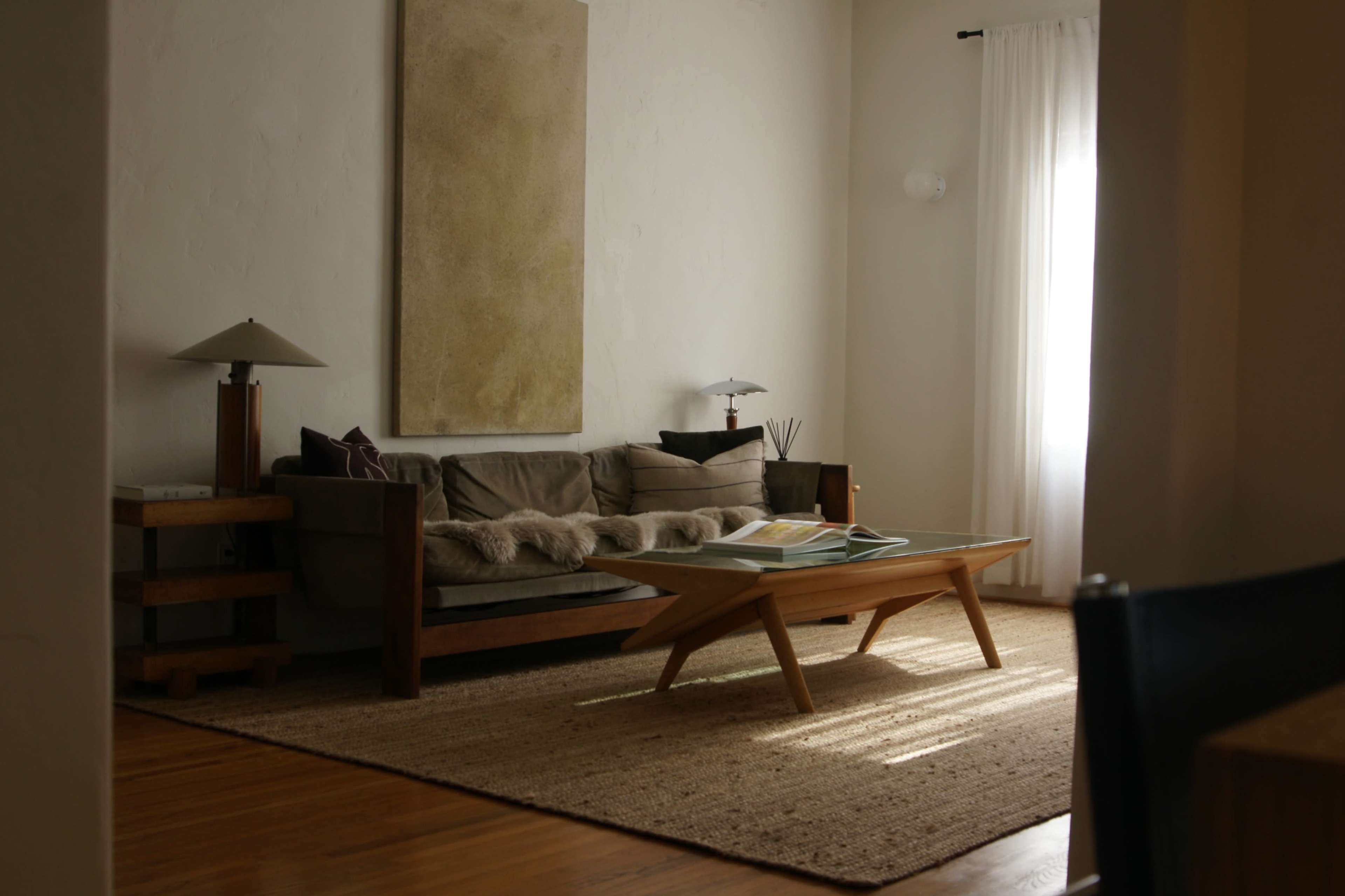 A minimalist living room features a wooden sofa, a coffee table, and a large wall art piece, with natural light streaming in through a nearby window.