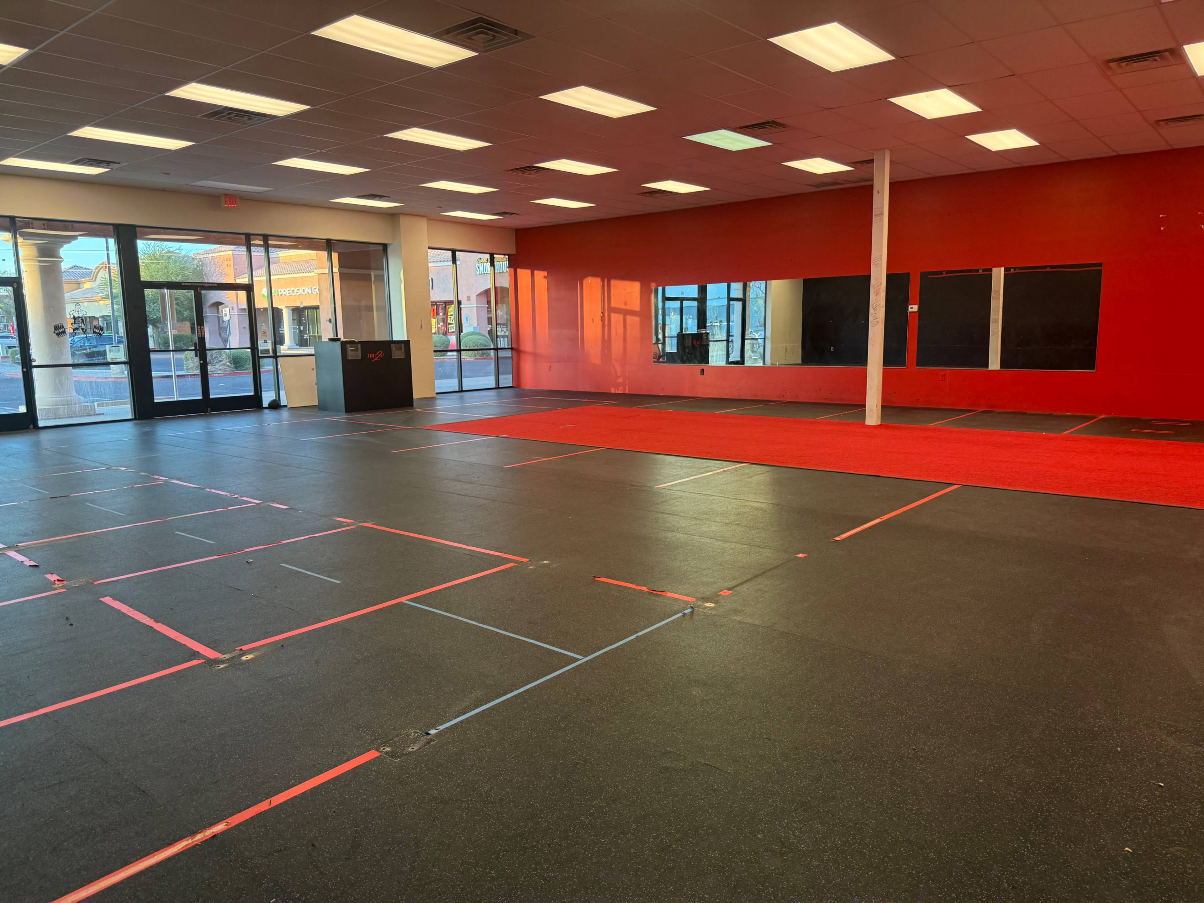 The image shows an empty fitness studio featuring black rubber flooring, red accent walls, and large windows allowing natural light to enter.