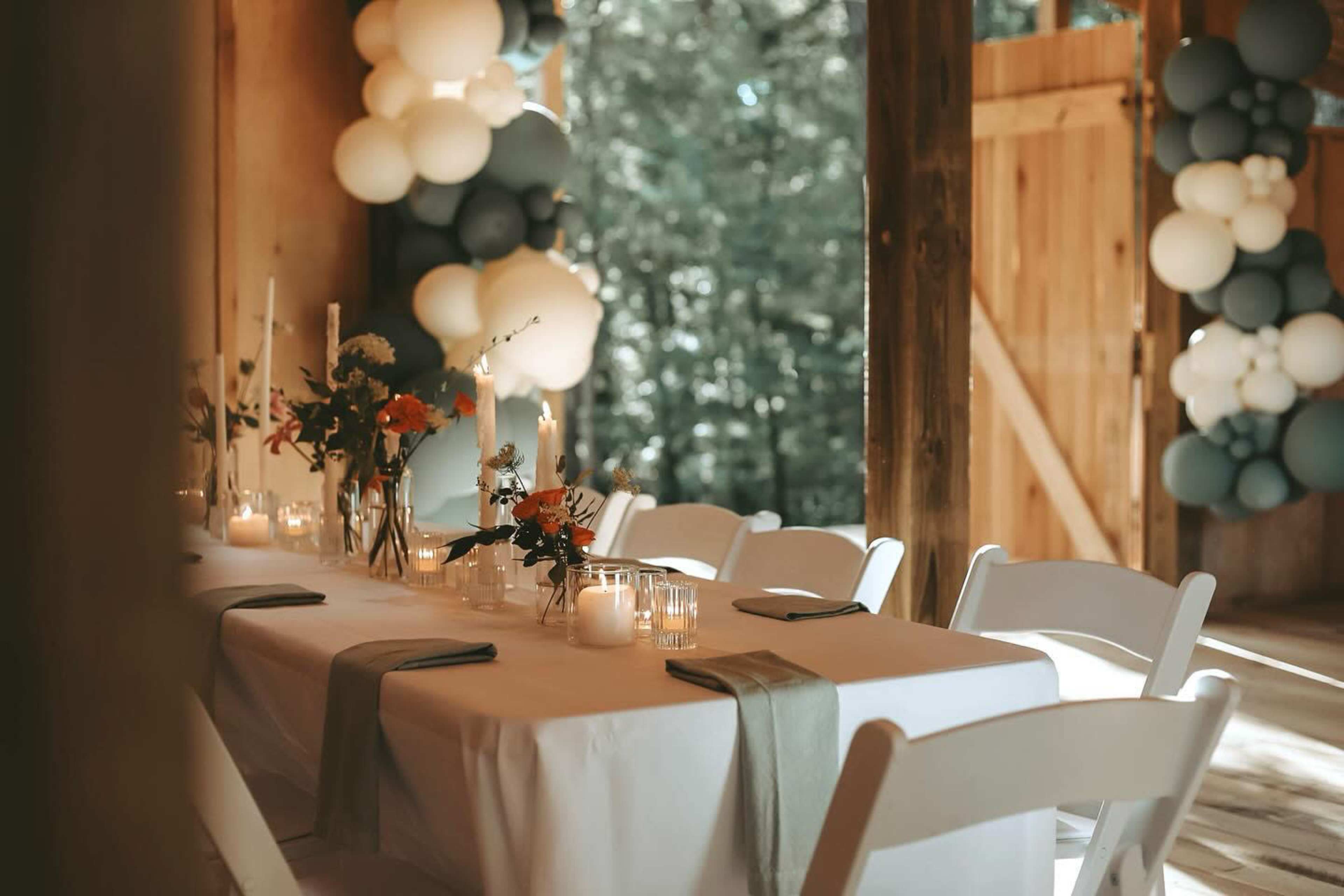 A decorated table is set up in a rustic space with floral centerpieces, candles, and a backdrop of balloon arrangements.
