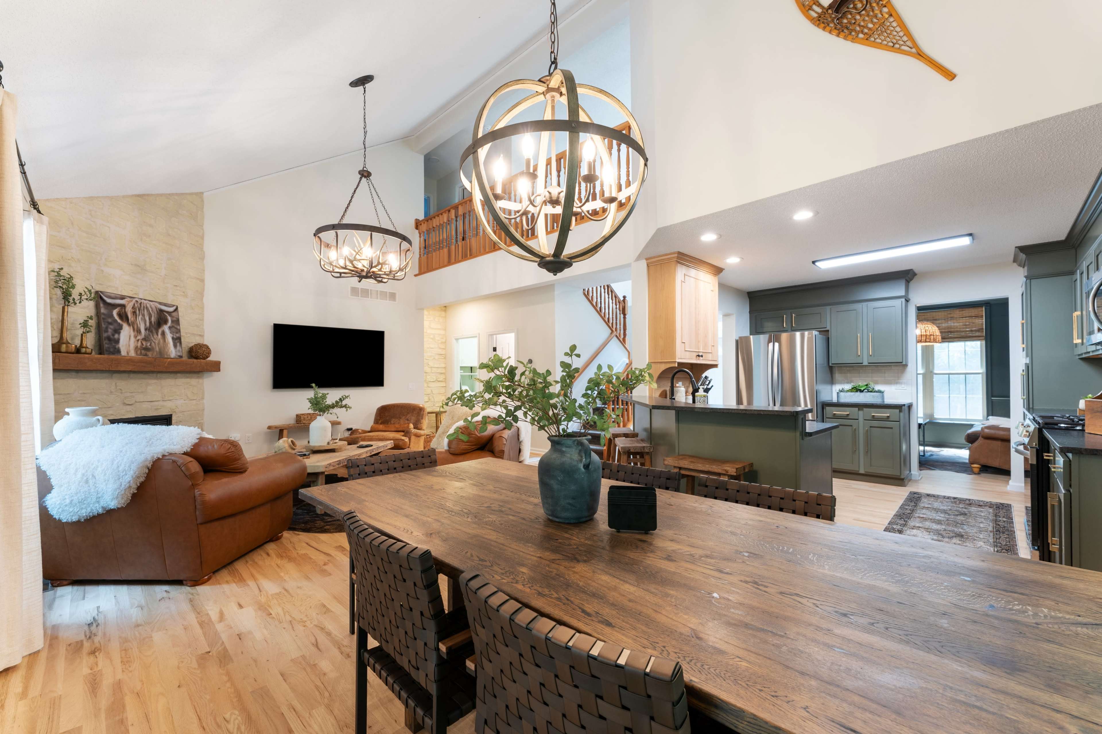The image shows a spacious modern living area with a dining table, leather seating, and a kitchen, all featuring light-colored wood and warm tones.