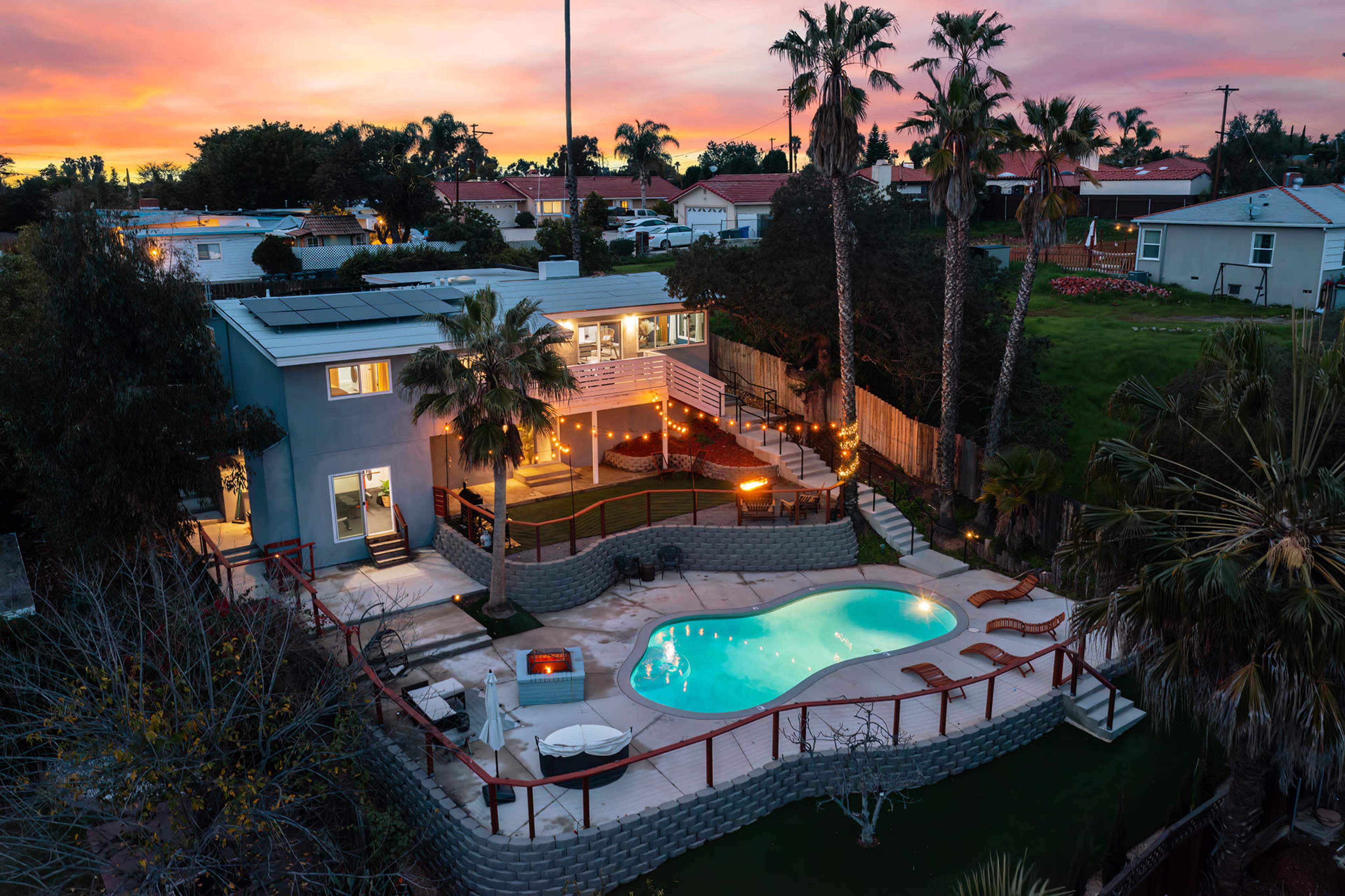 A modern house with a pool surrounded by palm trees, illuminated by lights at dusk.