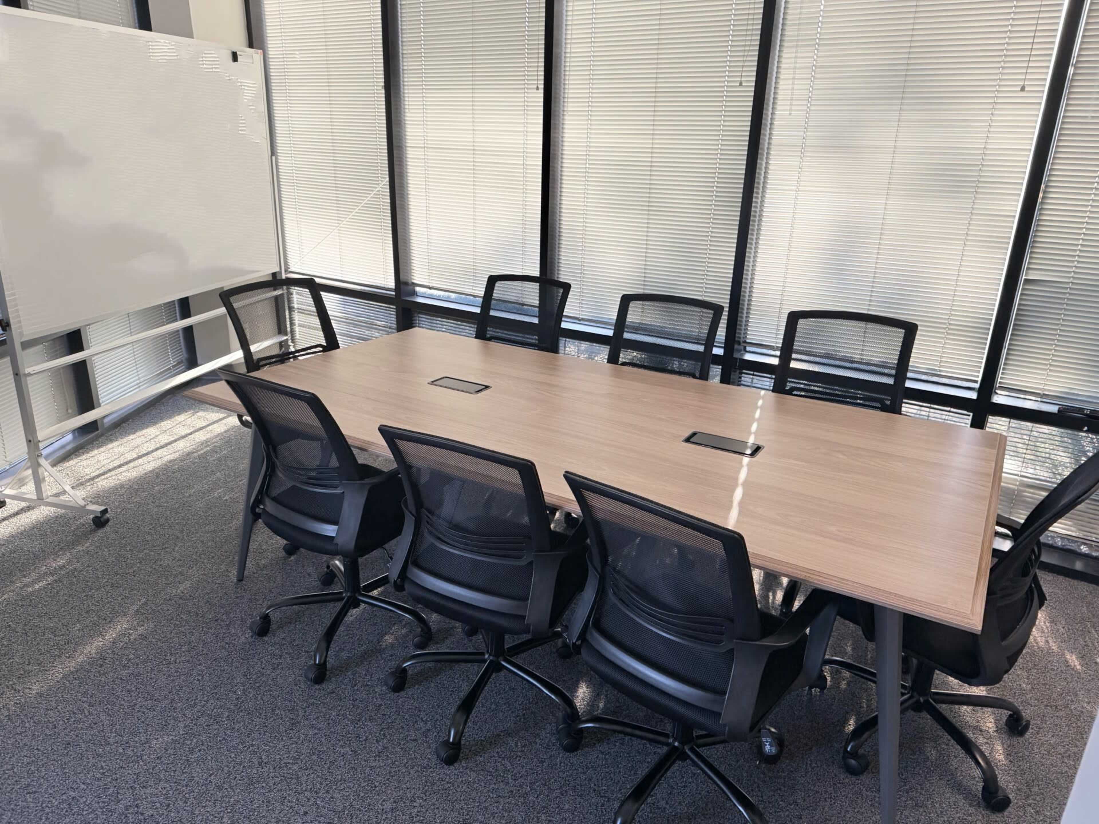 A modern conference room features a long wooden table surrounded by six black chairs, with a whiteboard positioned nearby and large windows letting in natural light.
