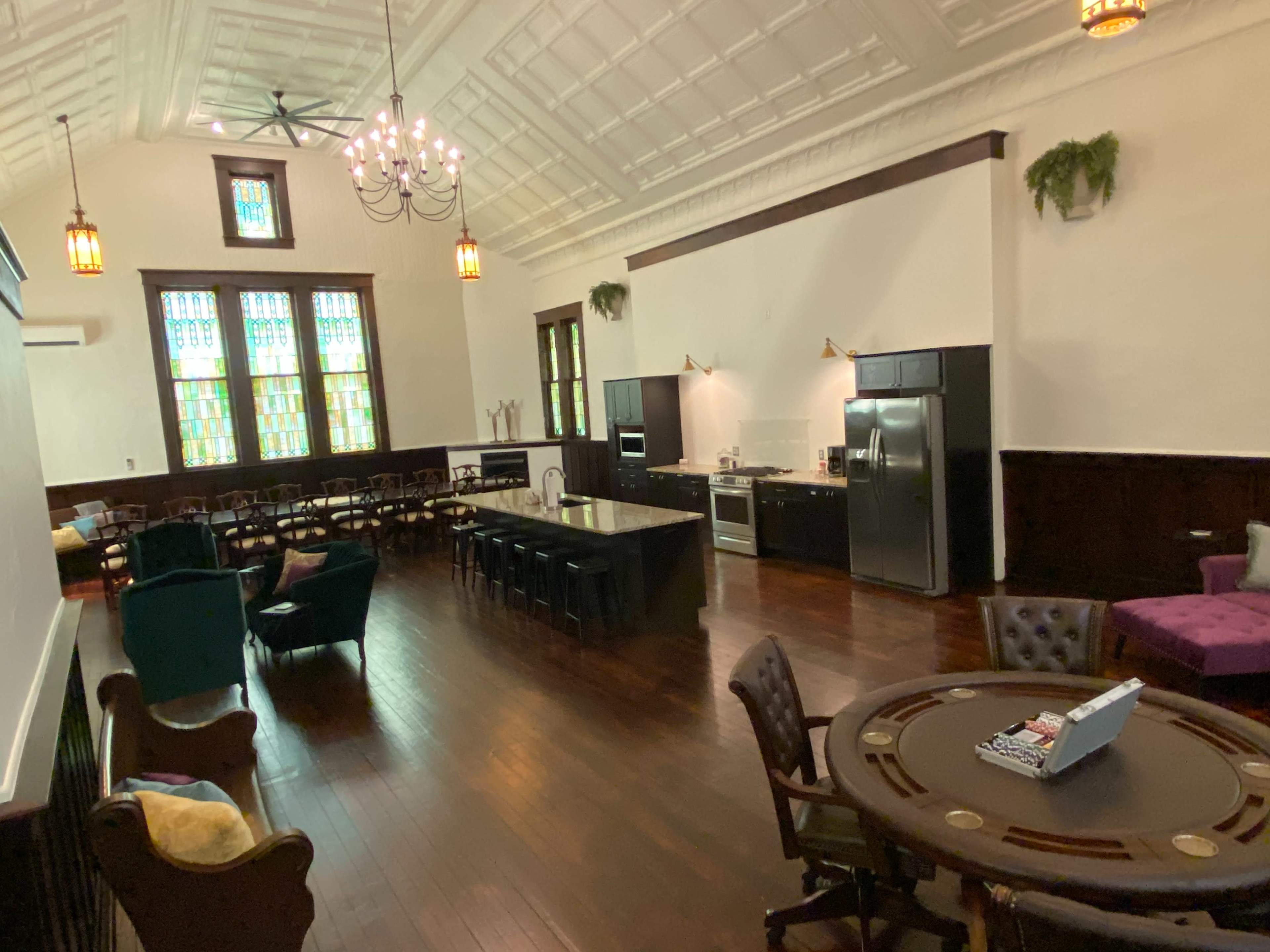 The image shows a spacious room with a high ceiling, featuring dining tables on one side and a kitchen area on the other, along with stained glass windows and wooden floors.