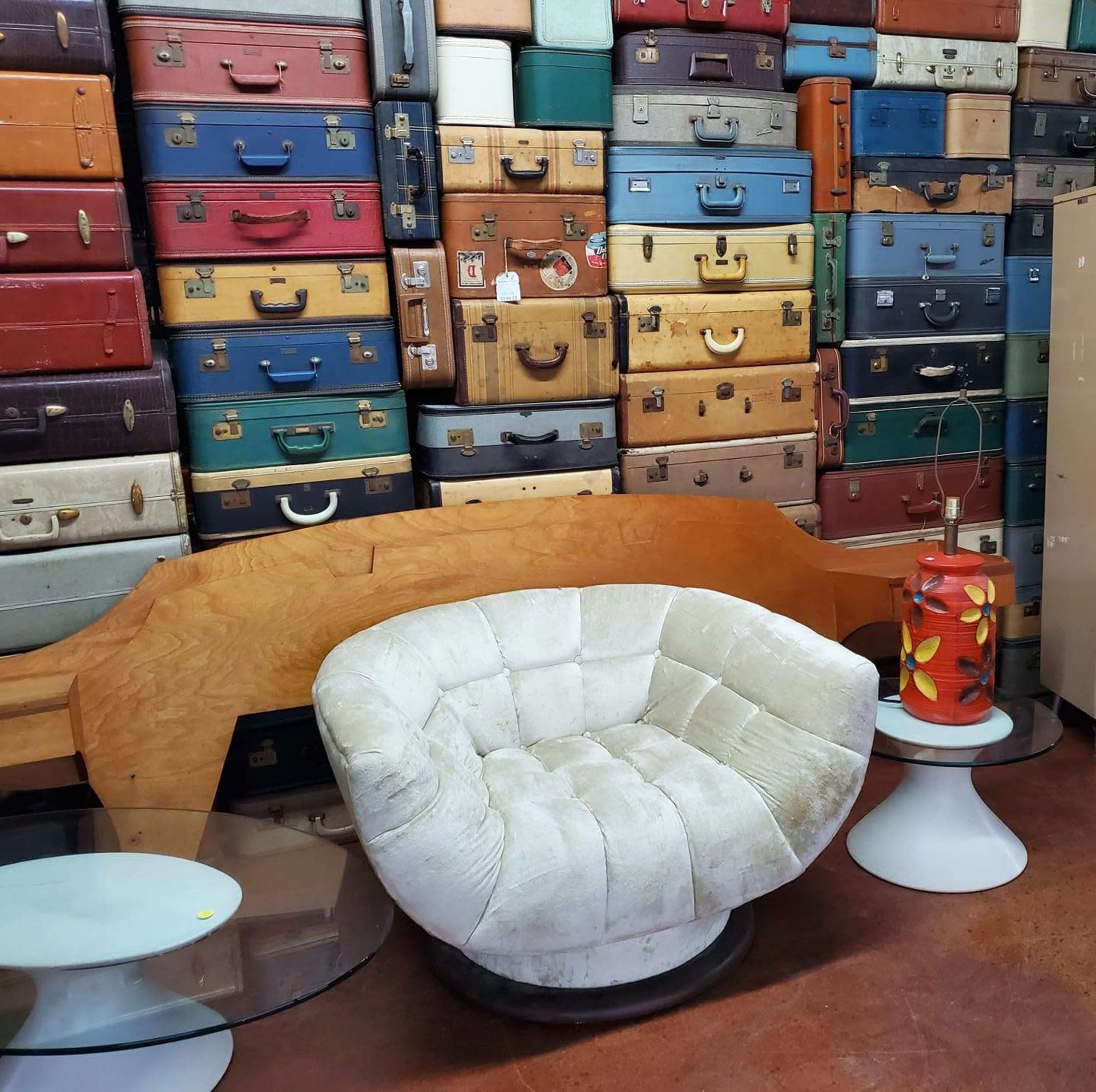 A white circular chair sits in front of a wall stacked with colorful vintage suitcases.
