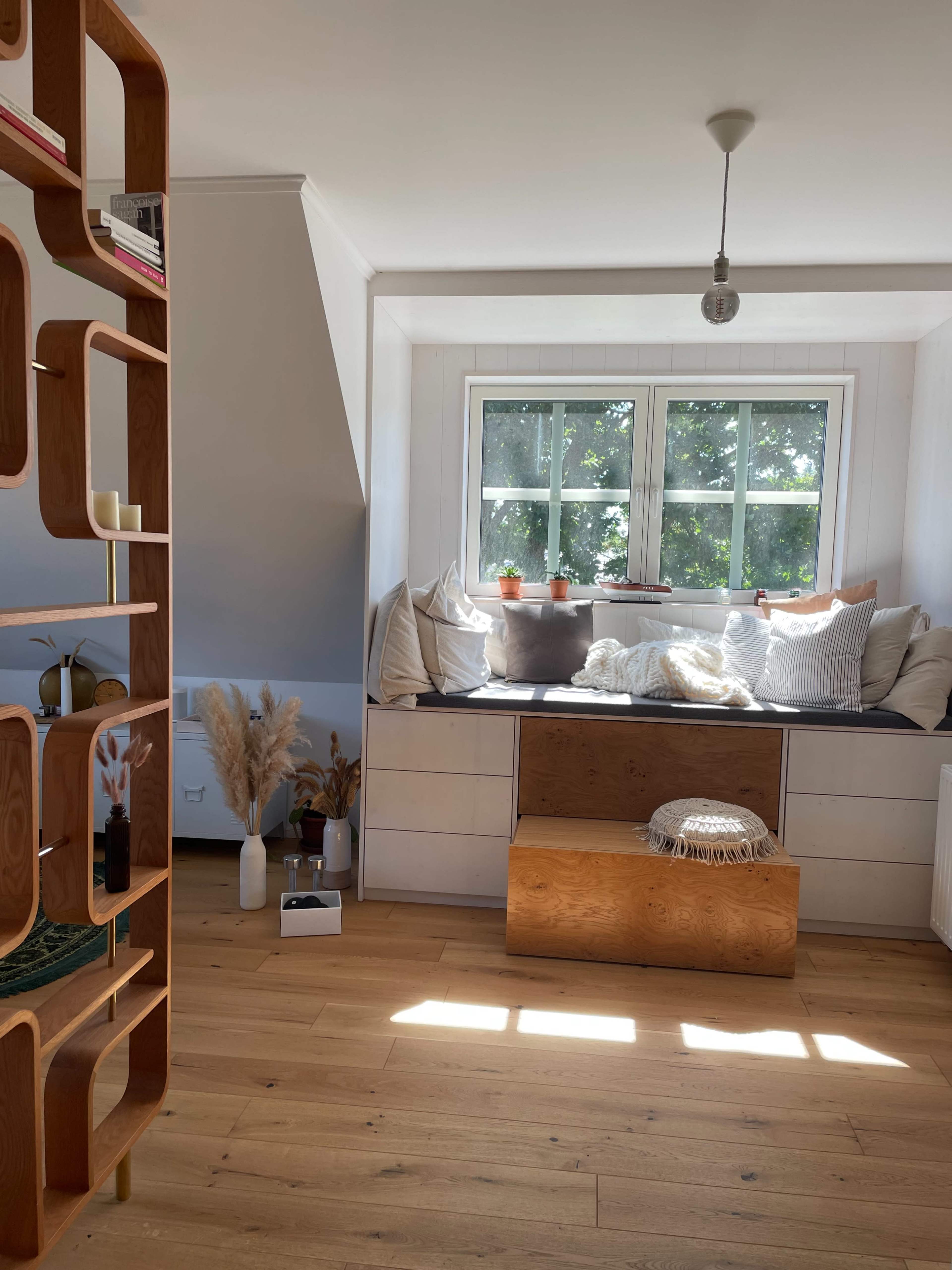 The image shows a bright room with a cozy window seat filled with cushions and plants, and a wooden bookshelf separating the space.