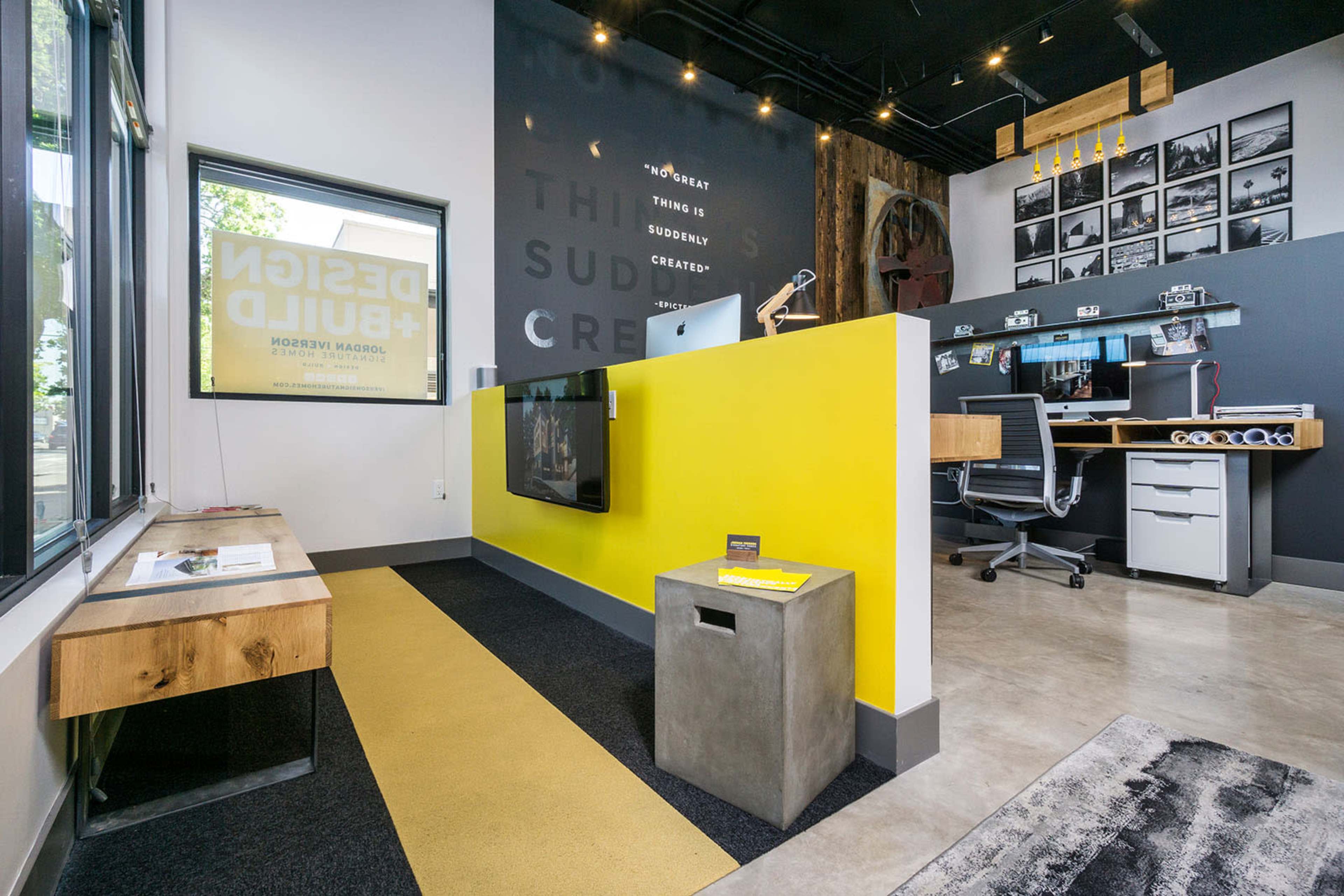 A modern office space featuring a yellow wall, a wooden bench, a concrete table, and a workspace with multiple desks and a large monitor.