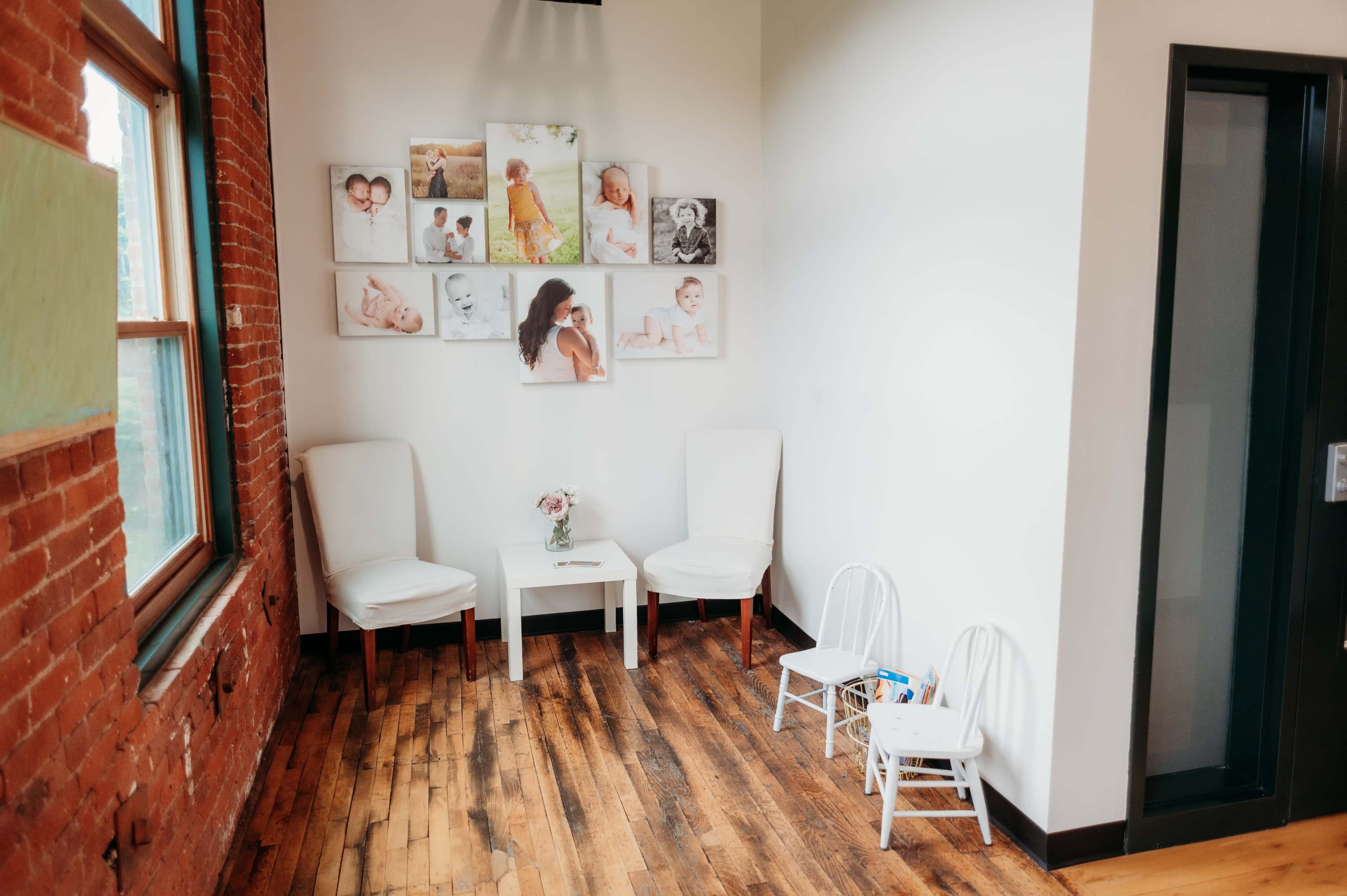 A small waiting area with two white chairs and a table, featuring framed photographs on the wall and wooden flooring.