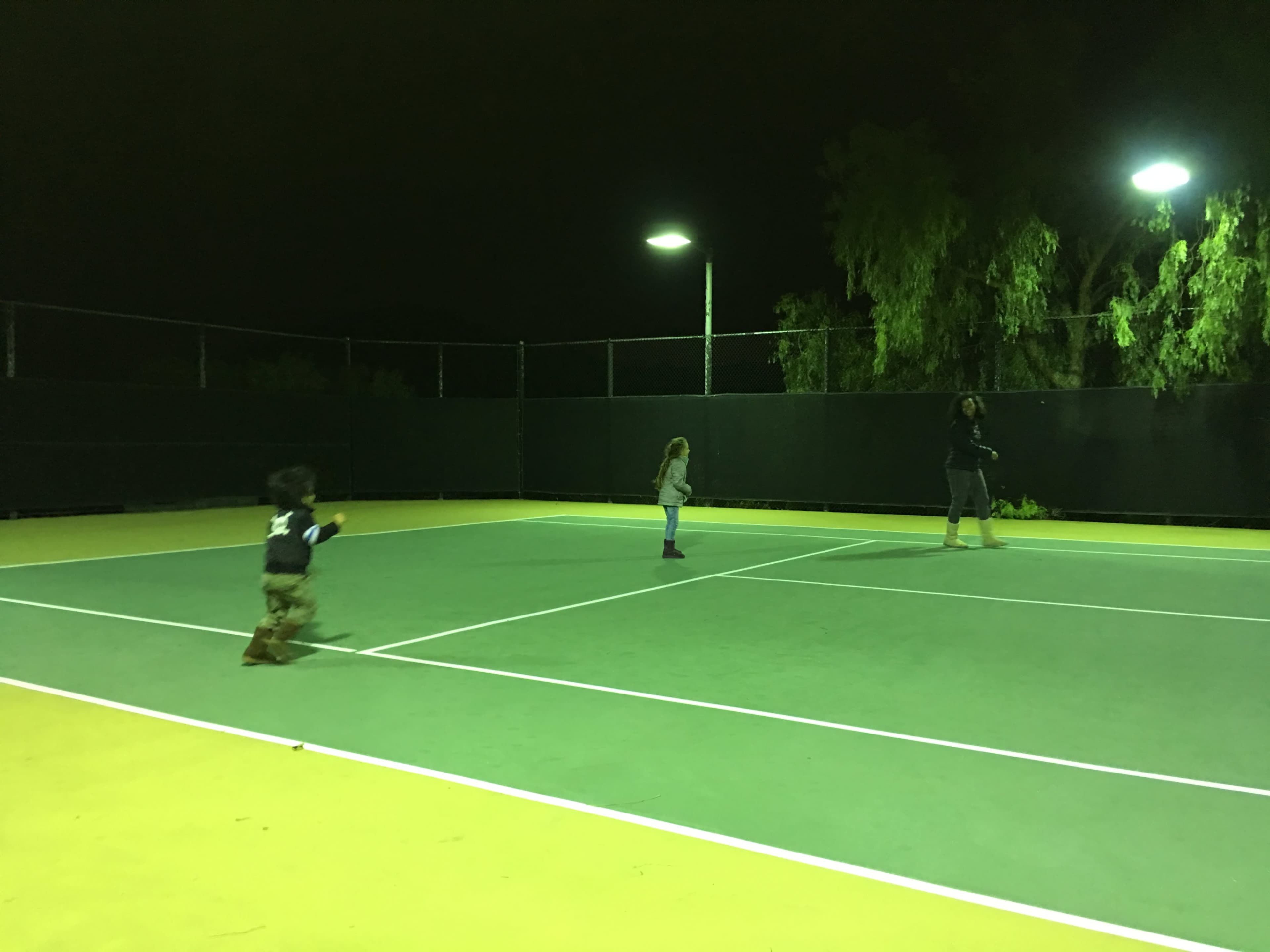 Two children play on a tennis court at night while an adult stands on the opposite side.