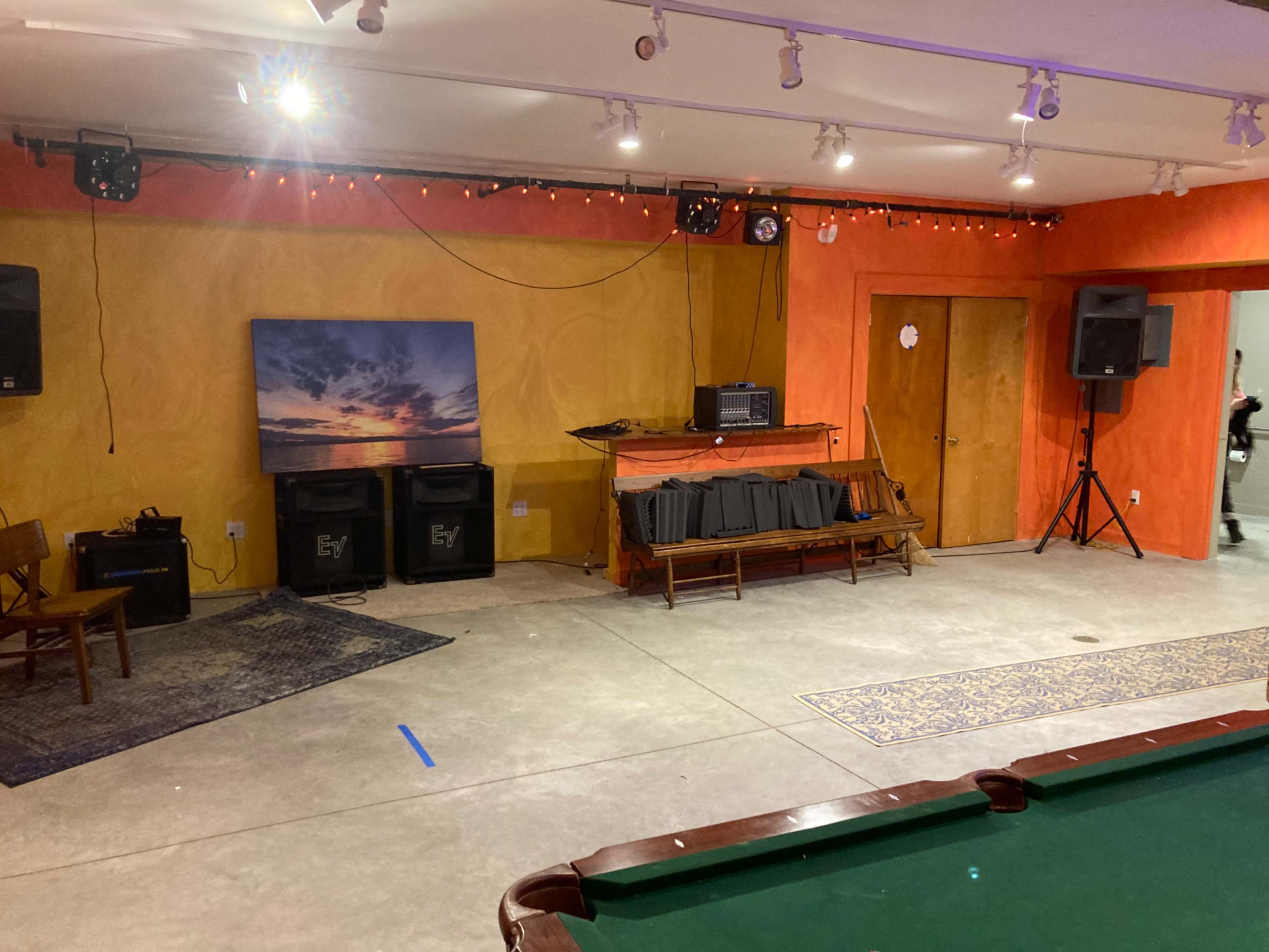 A room with a pool table, a wooden chair, sound equipment, and a wall-mounted television displaying a landscape scene, all under decorative lighting.
