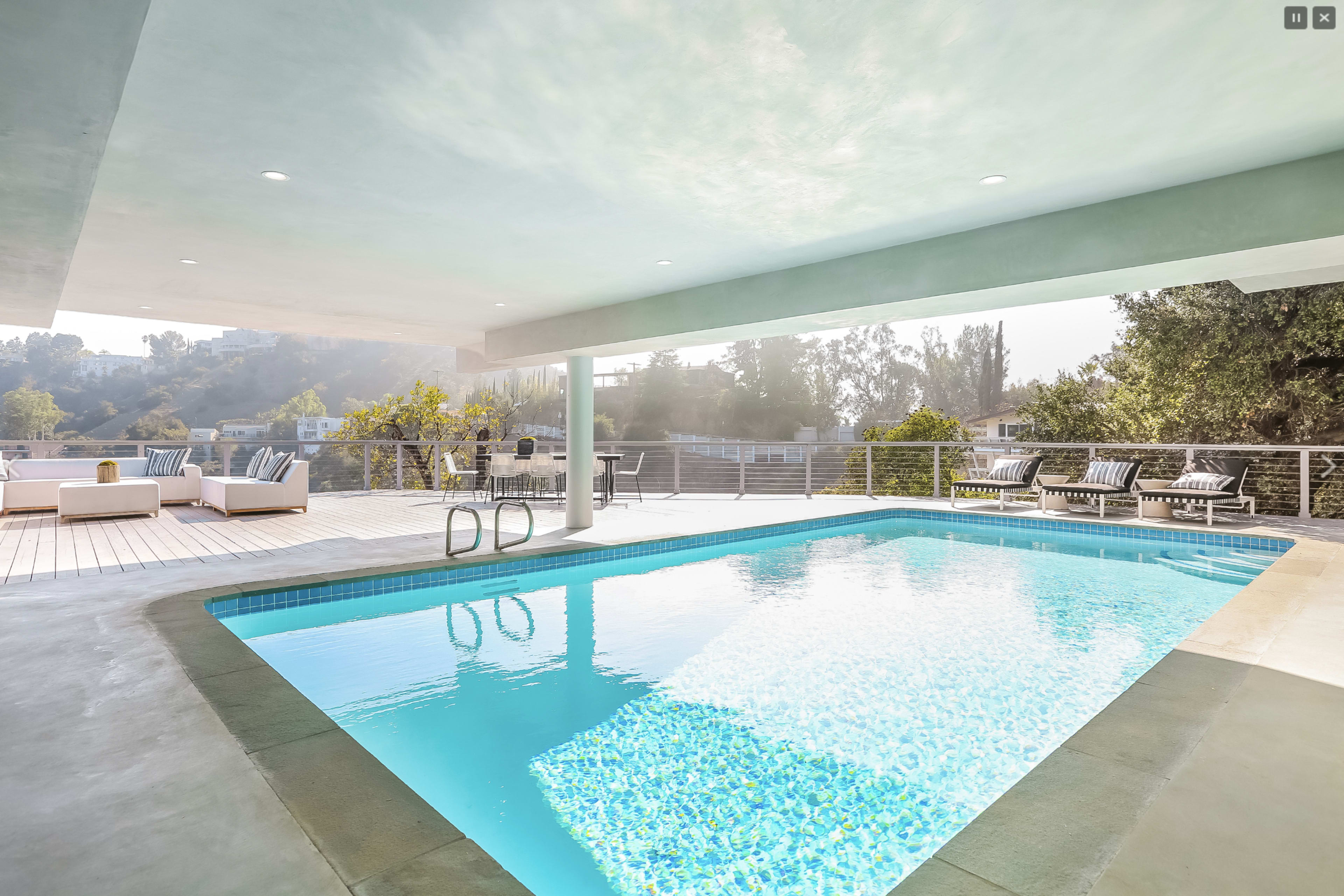 A modern outdoor area features a rectangular swimming pool surrounded by loungers and seating under a covered patio.