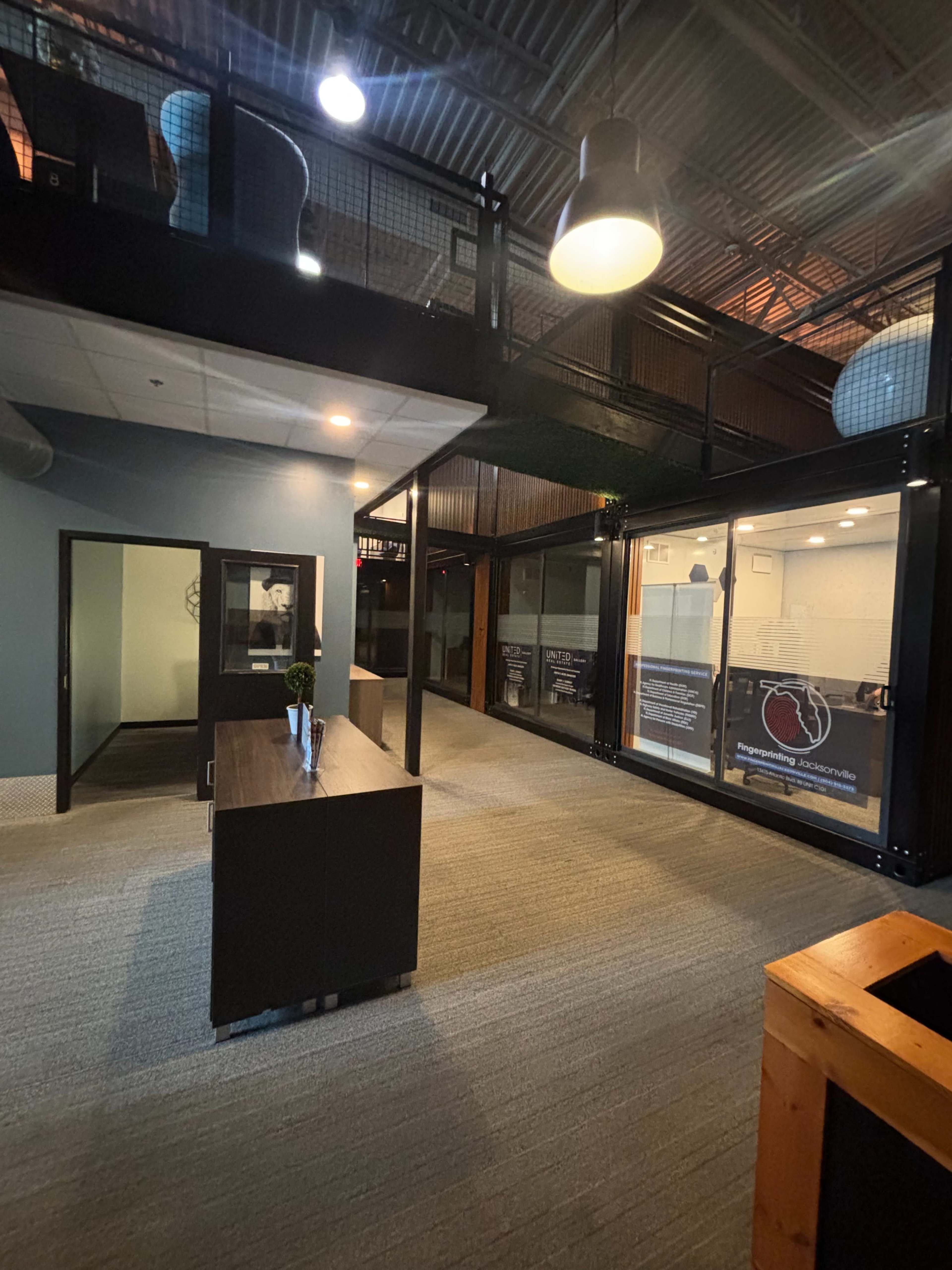 The image shows a modern office space with a reception area, glass-walled meeting rooms, and an upper level visible in the background.