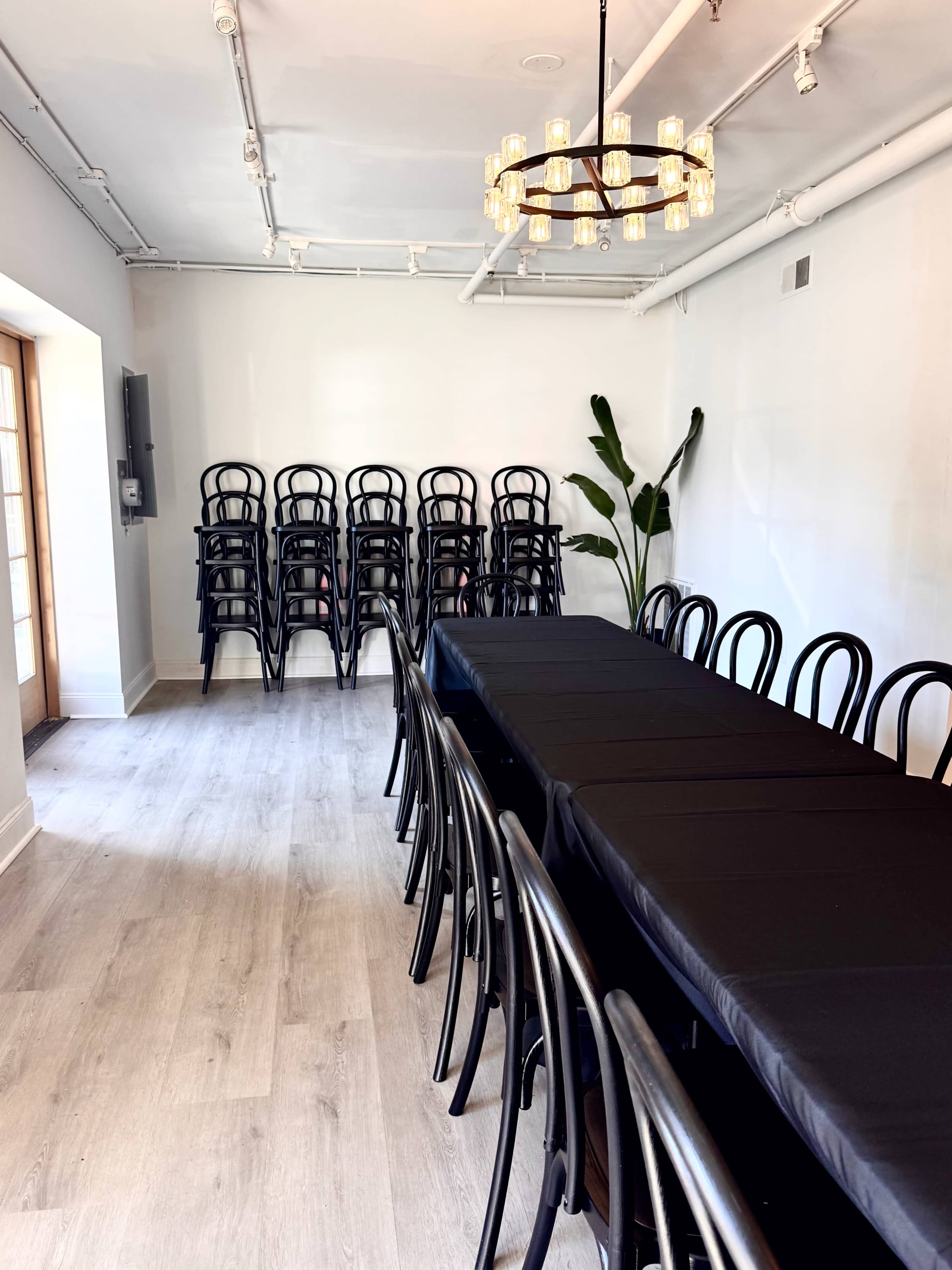 The image shows a rectangular dining table covered with a black cloth, surrounded by black chairs, in a room with a chandelier and a stack of chairs against one wall.