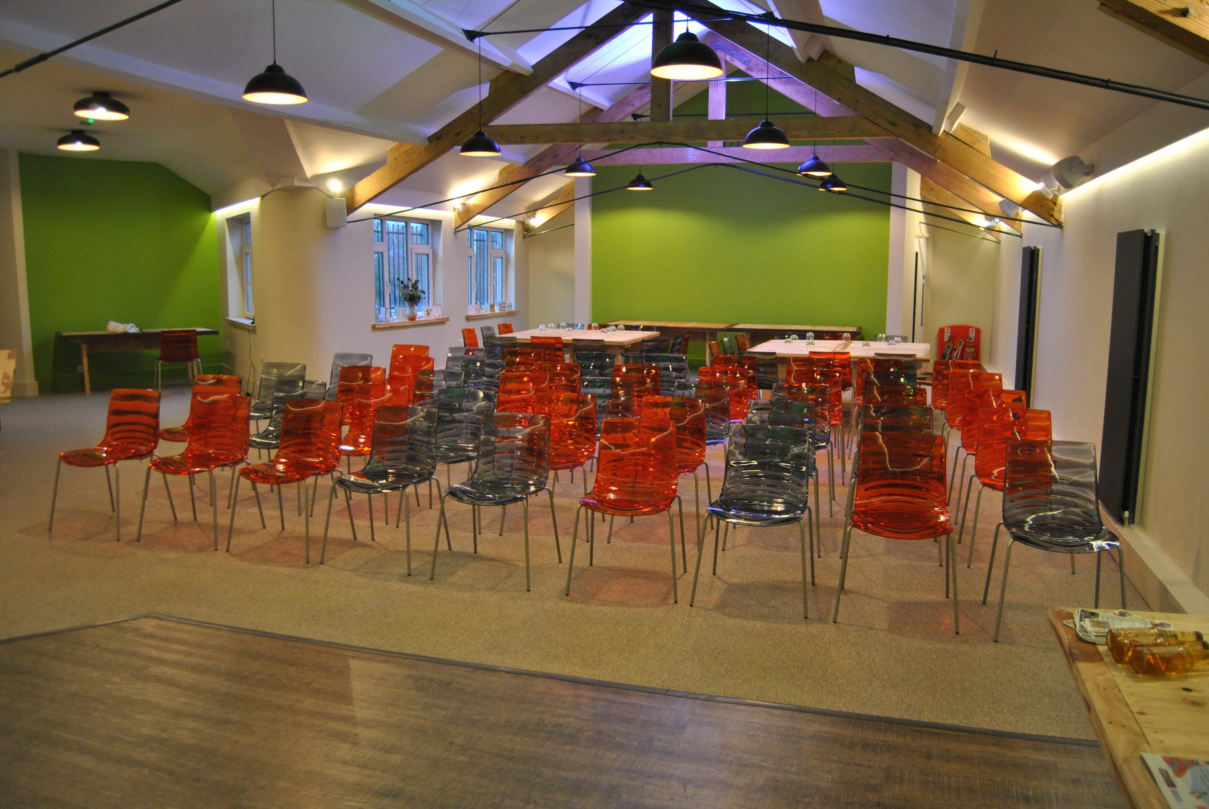 A room with a high ceiling features rows of red and gray transparent chairs arranged for an event, with green walls and wooden beams visible above.
