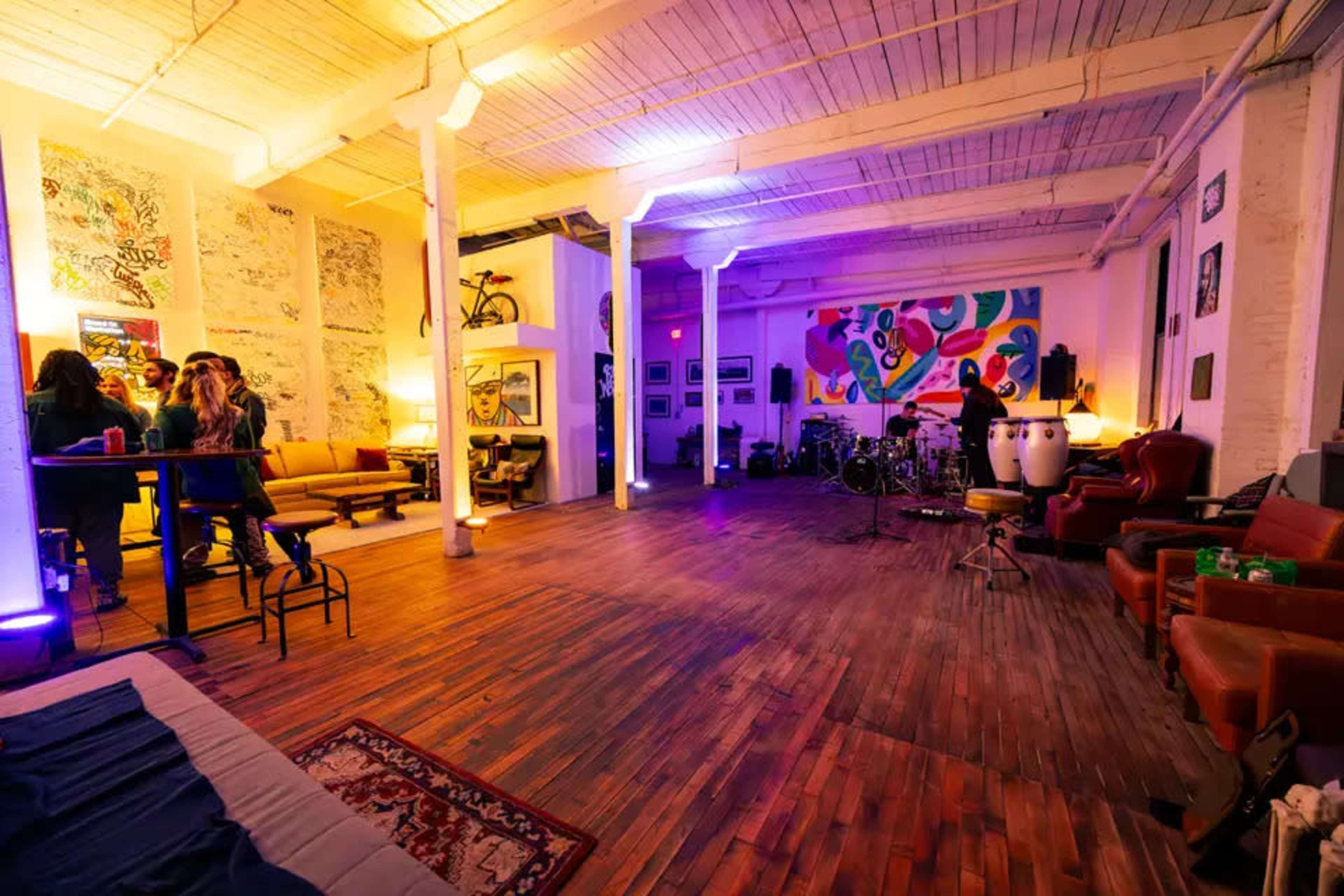 The image shows a spacious, illuminated room with a wooden floor, featuring colorful wall art, seating areas, and musical instruments setup.