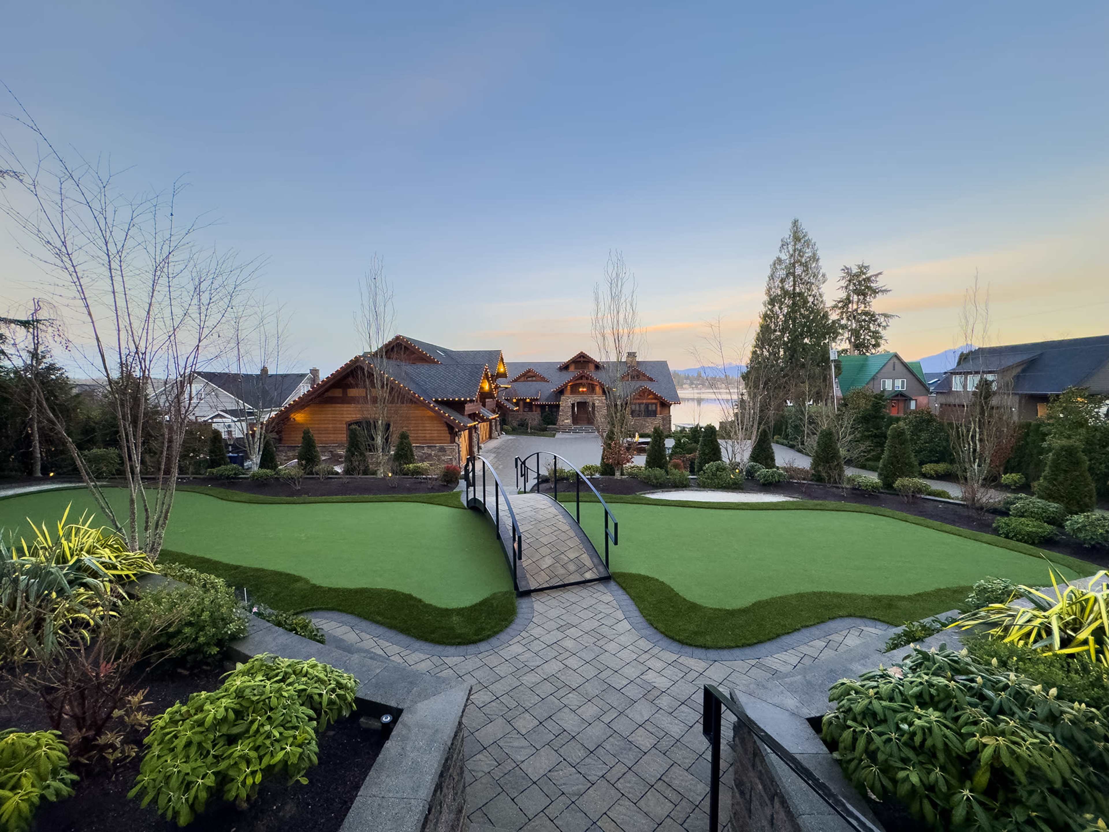 A pathway with a bridge connects manicured lawns to a wooden lodge in a scenic landscape.