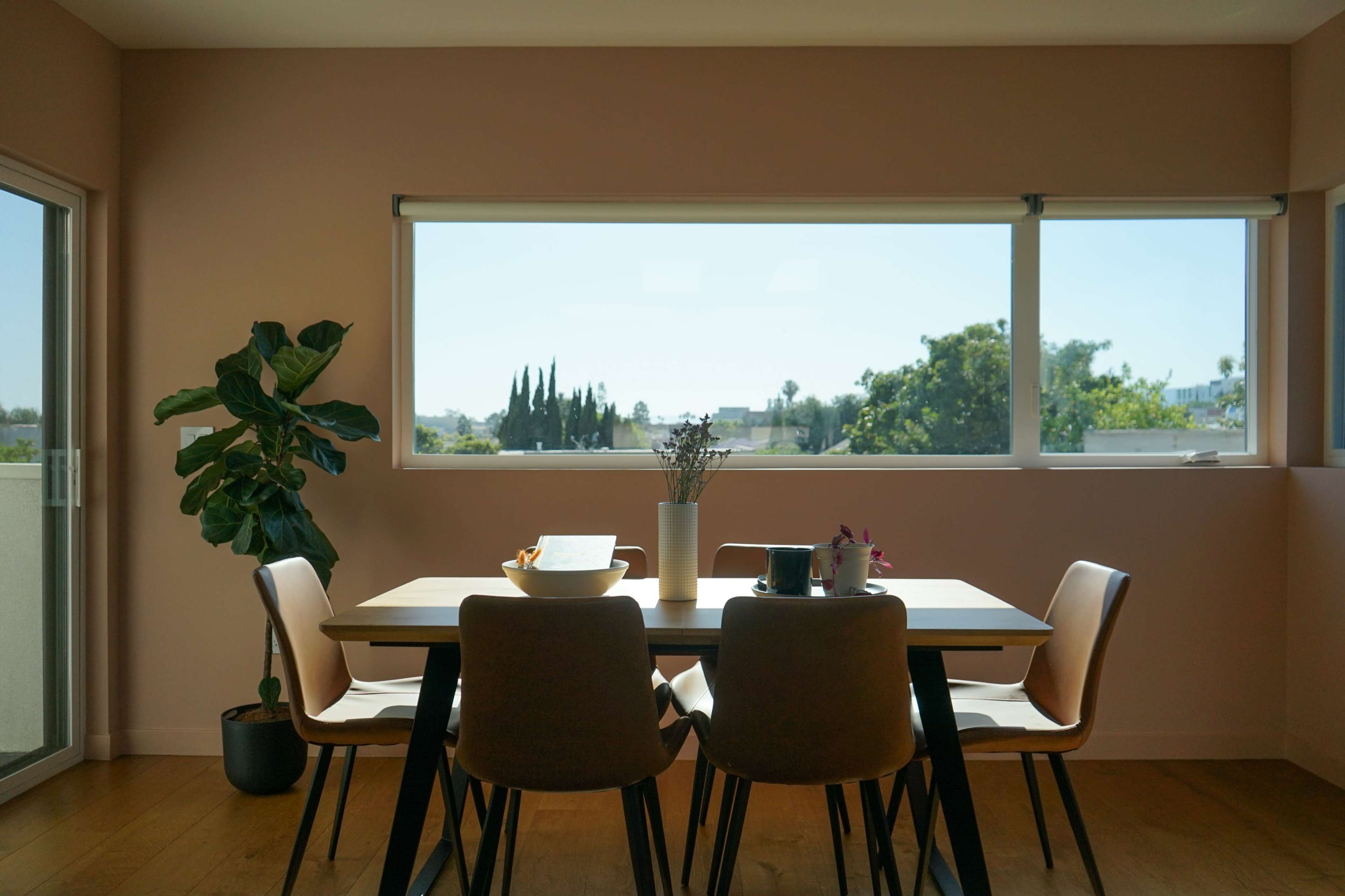 A dining table with chairs is positioned near a large window, offering a view of greenery outside.