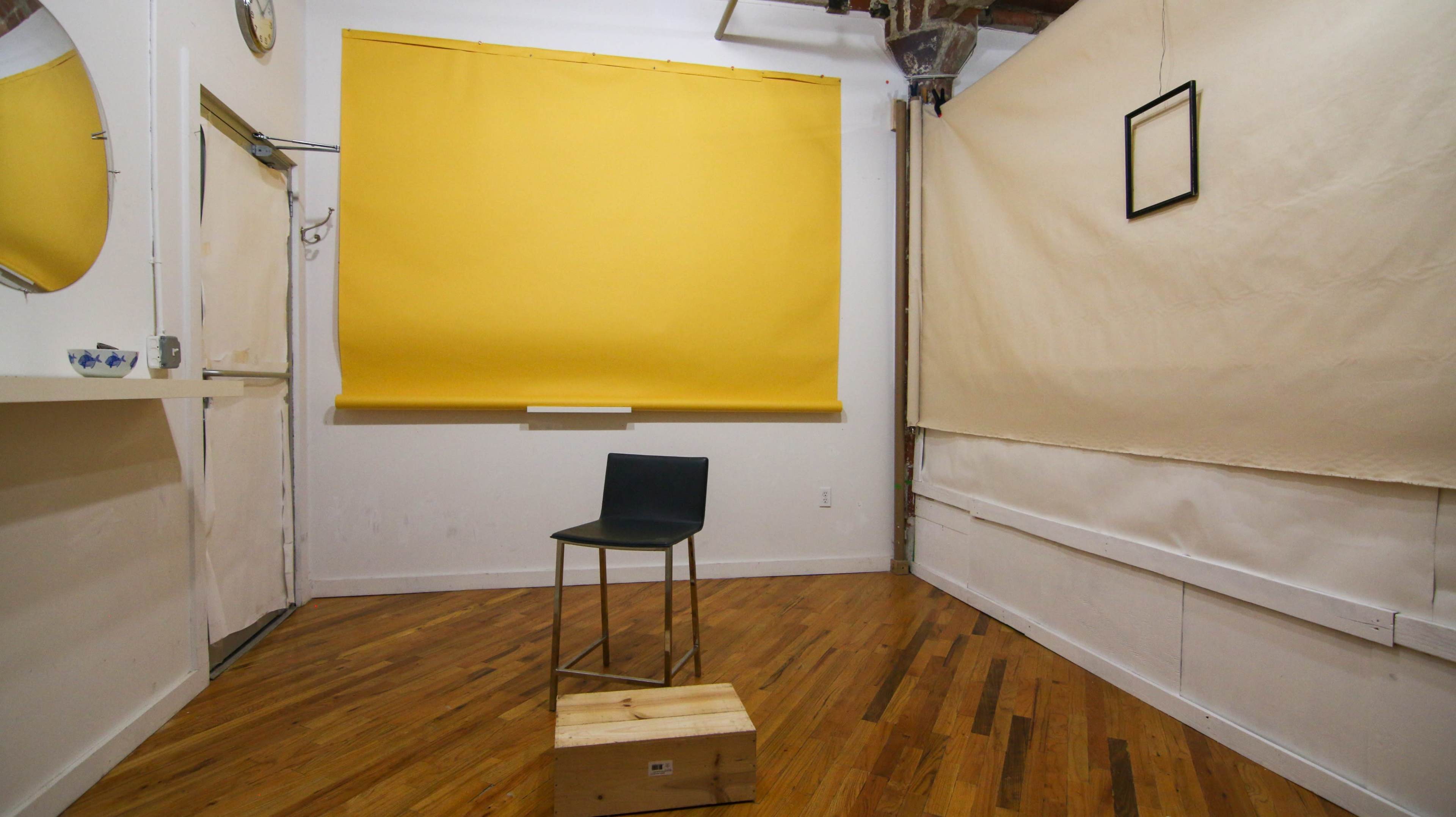 The image shows a small room with wooden flooring, featuring a yellow wall and a black chair on a wooden box positioned in the center.