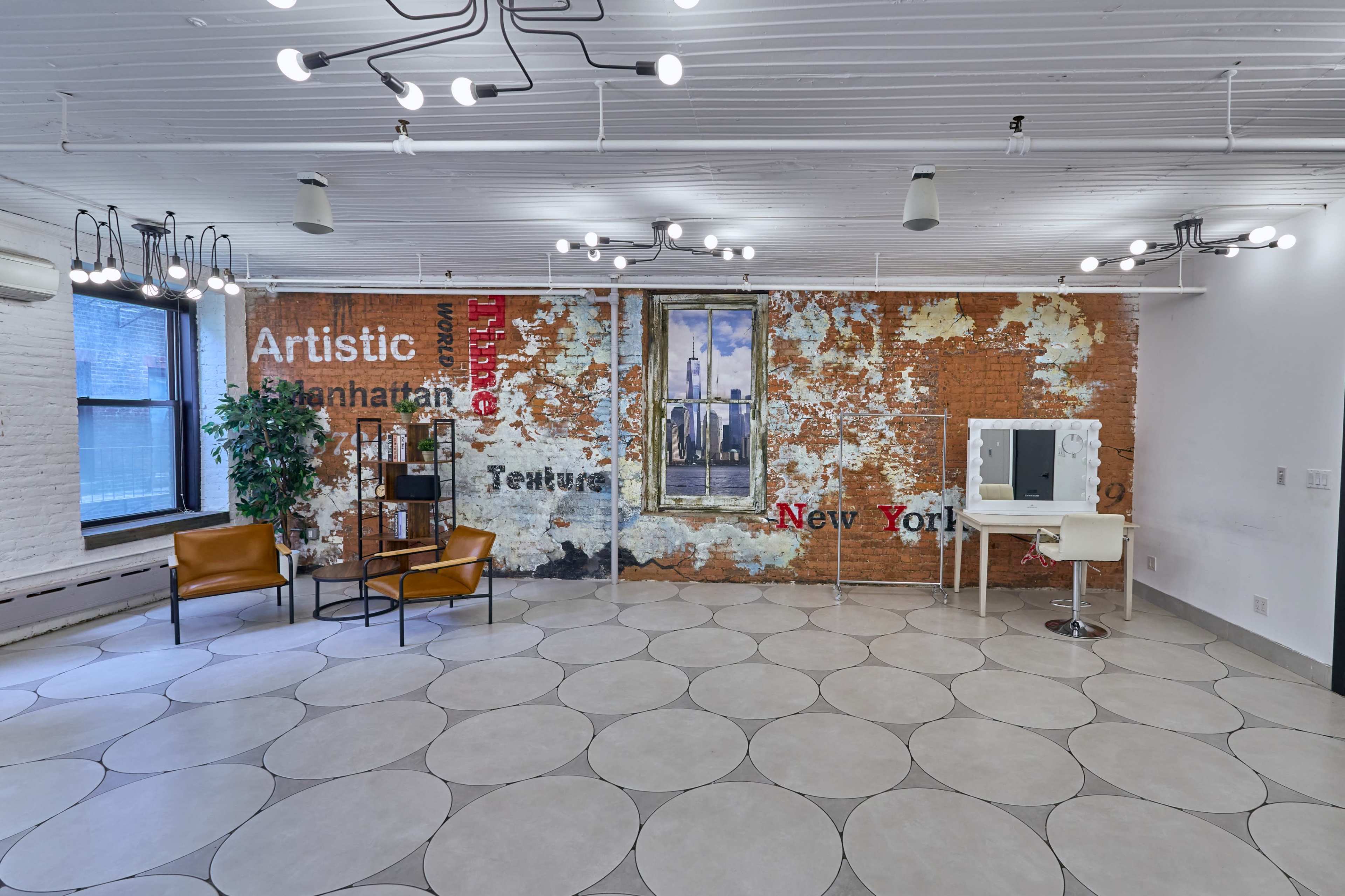 An empty room with a brick wall displaying artistic text and a painting, along with a circular tile floor and minimalist furniture consisting of two chairs and a vanity.