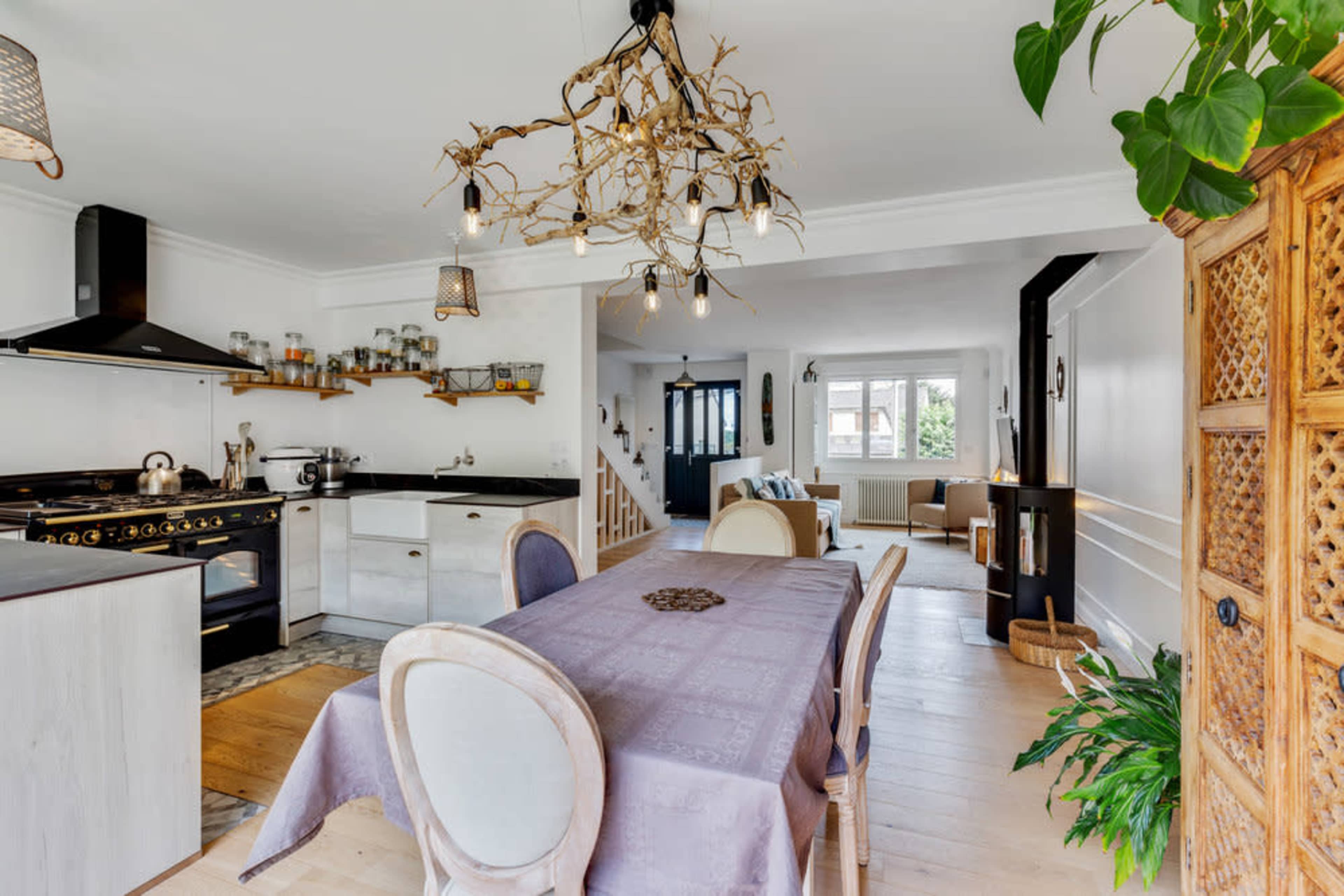 A modern kitchen and dining area features a wooden table with chairs, an ornate light fixture, and a view into a living space with natural light.