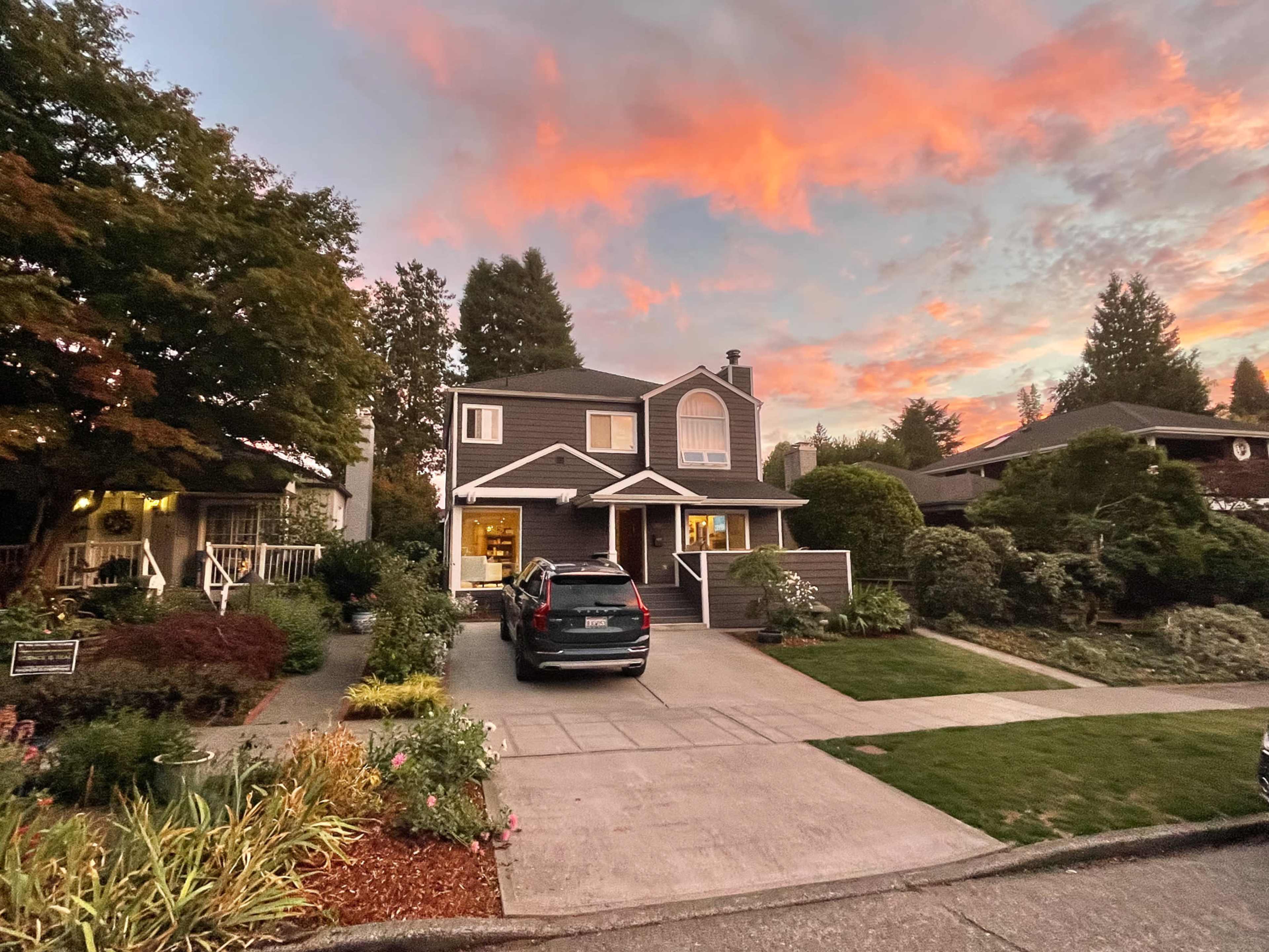 A two-story house with a driveway is set against a colorful sunset sky, surrounded by well-maintained gardens and trees.