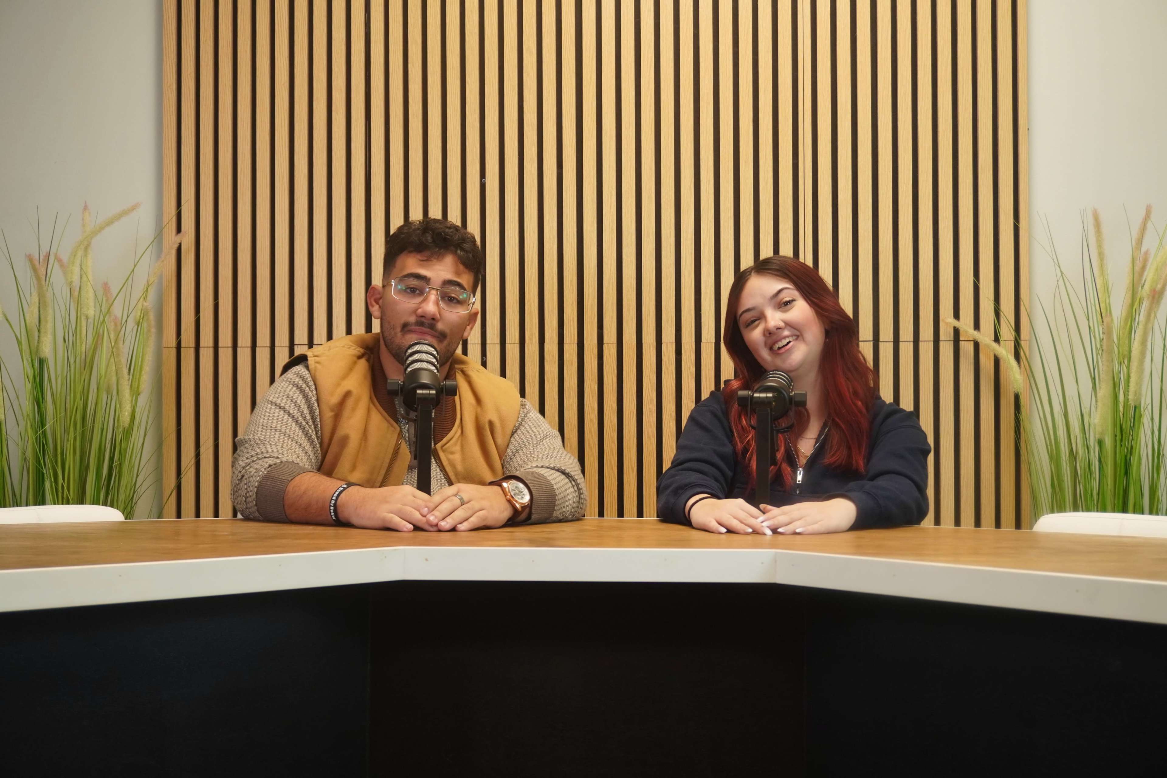 Two people sit at a table with microphones in front of them, surrounded by potted plants and a wooden wall.