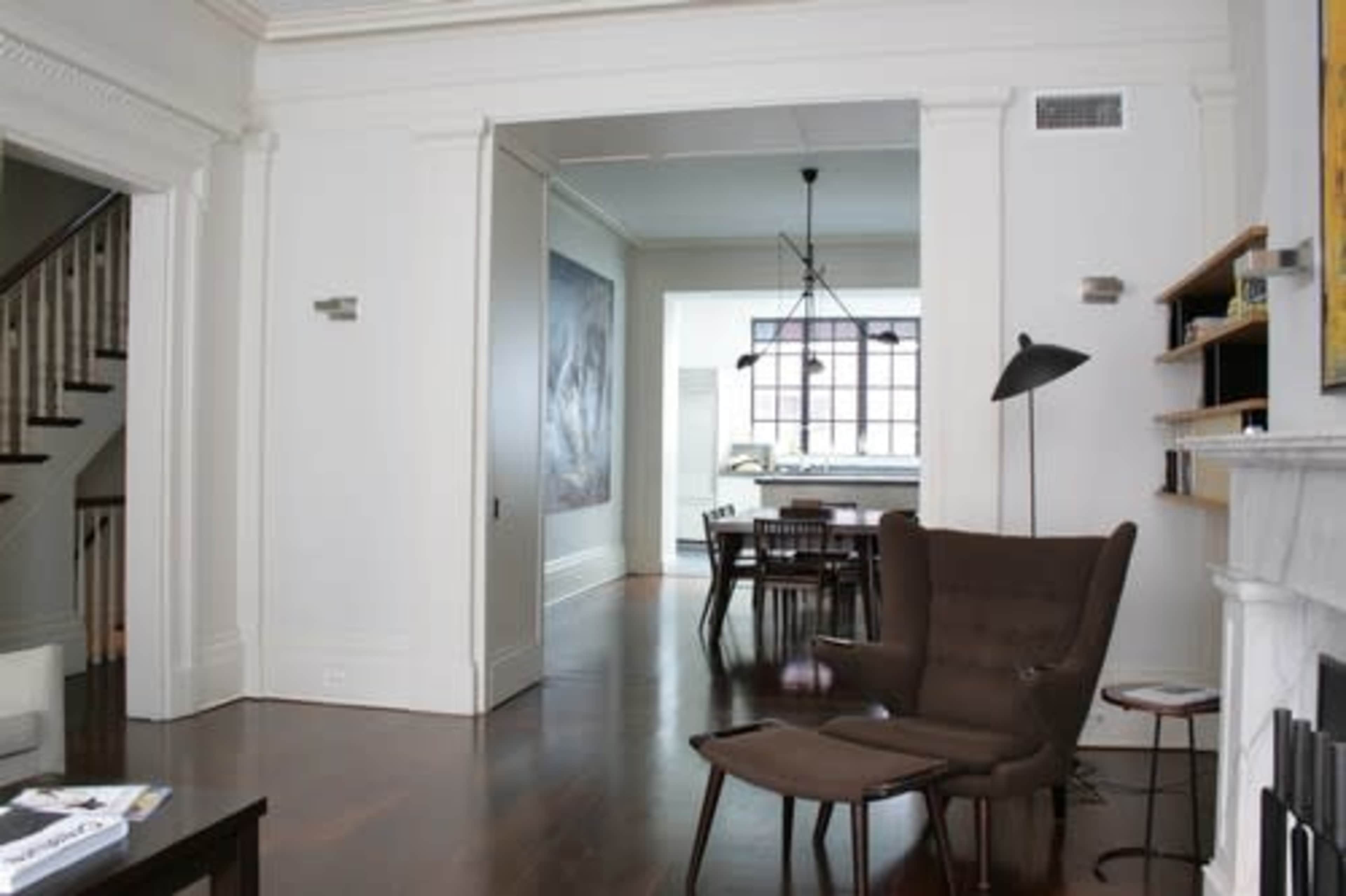 A spacious living area with a modern armchair, an open doorway leading to a dining area, and large windows illuminating the space.