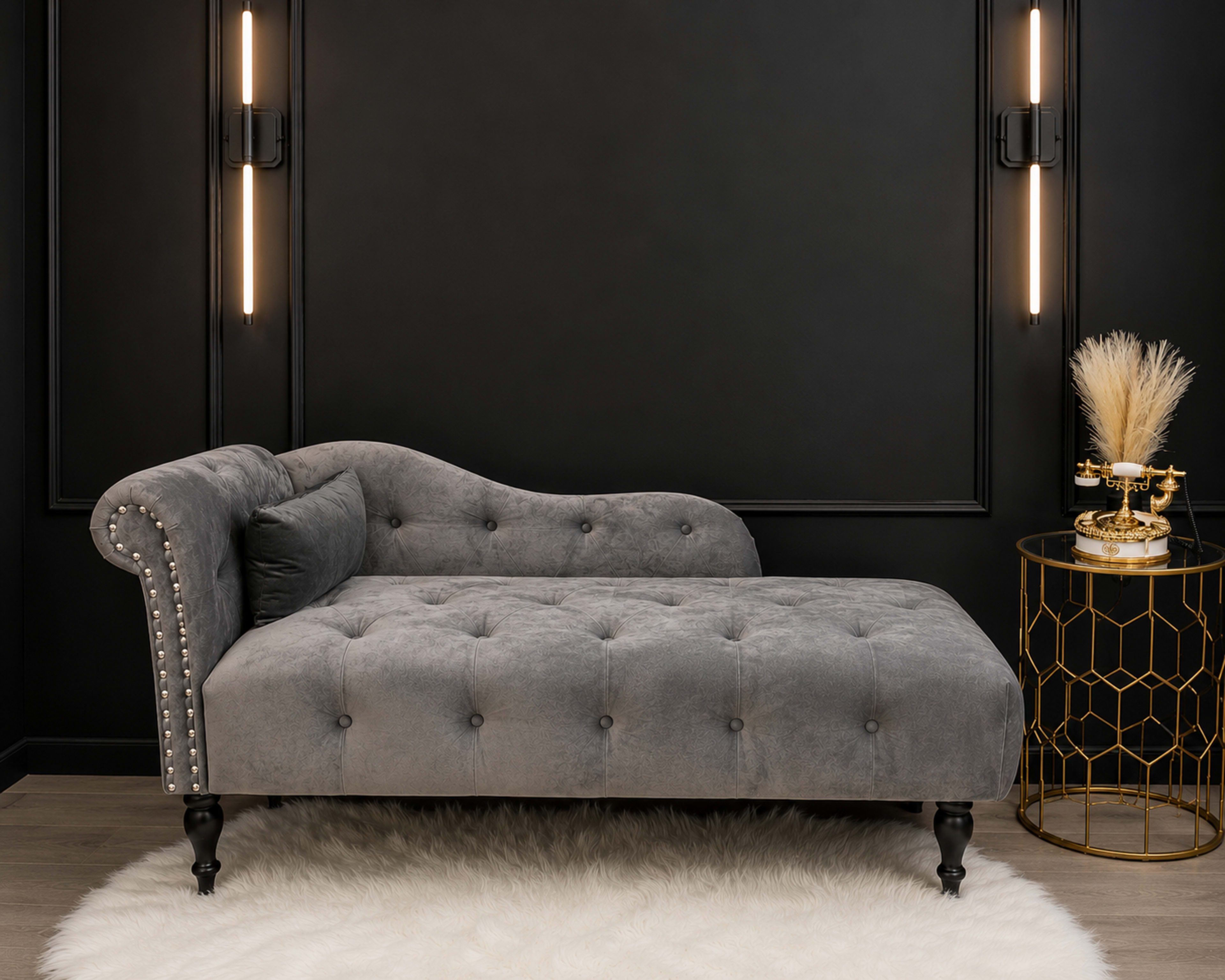 The image features a gray tufted chaise lounge placed against a black wall, accompanied by a gold side table with decorative elements and a plush white rug underneath.