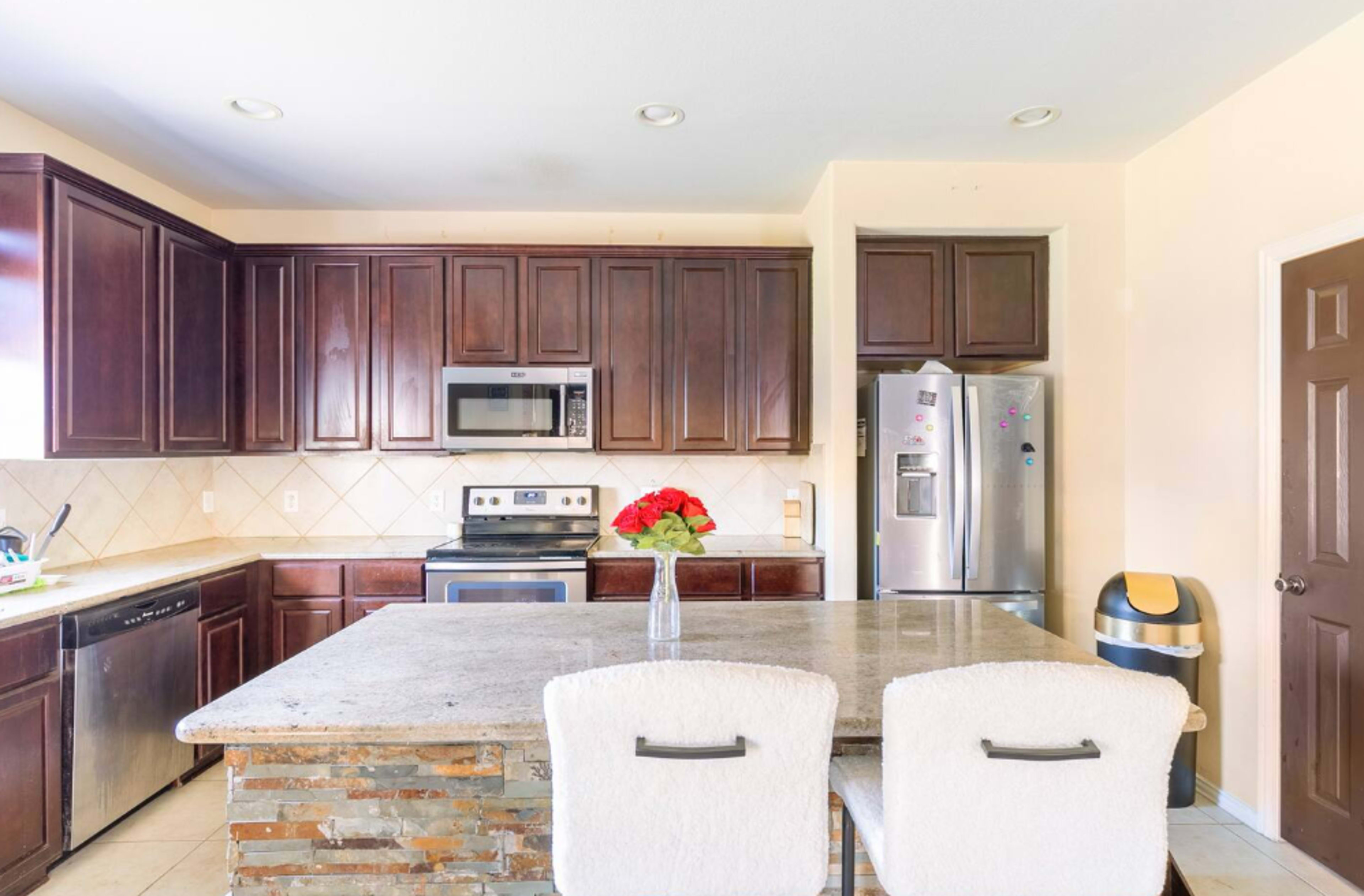 A modern kitchen with dark wood cabinets, stainless steel appliances, and a stone-topped island with two white chairs.