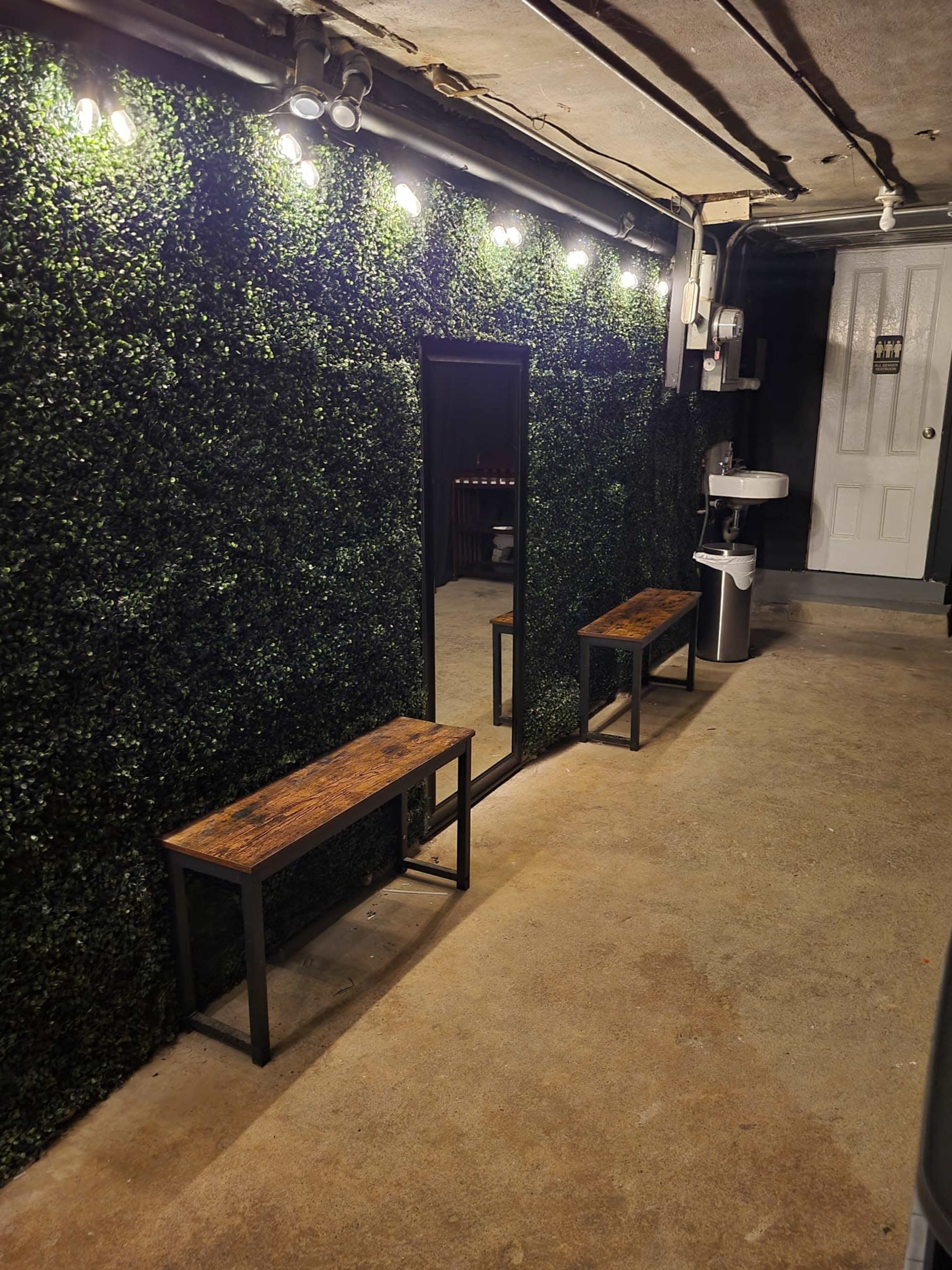 The image shows a basement room with a green artificial plant wall, two wooden benches, a mirror, and a small sink area.
