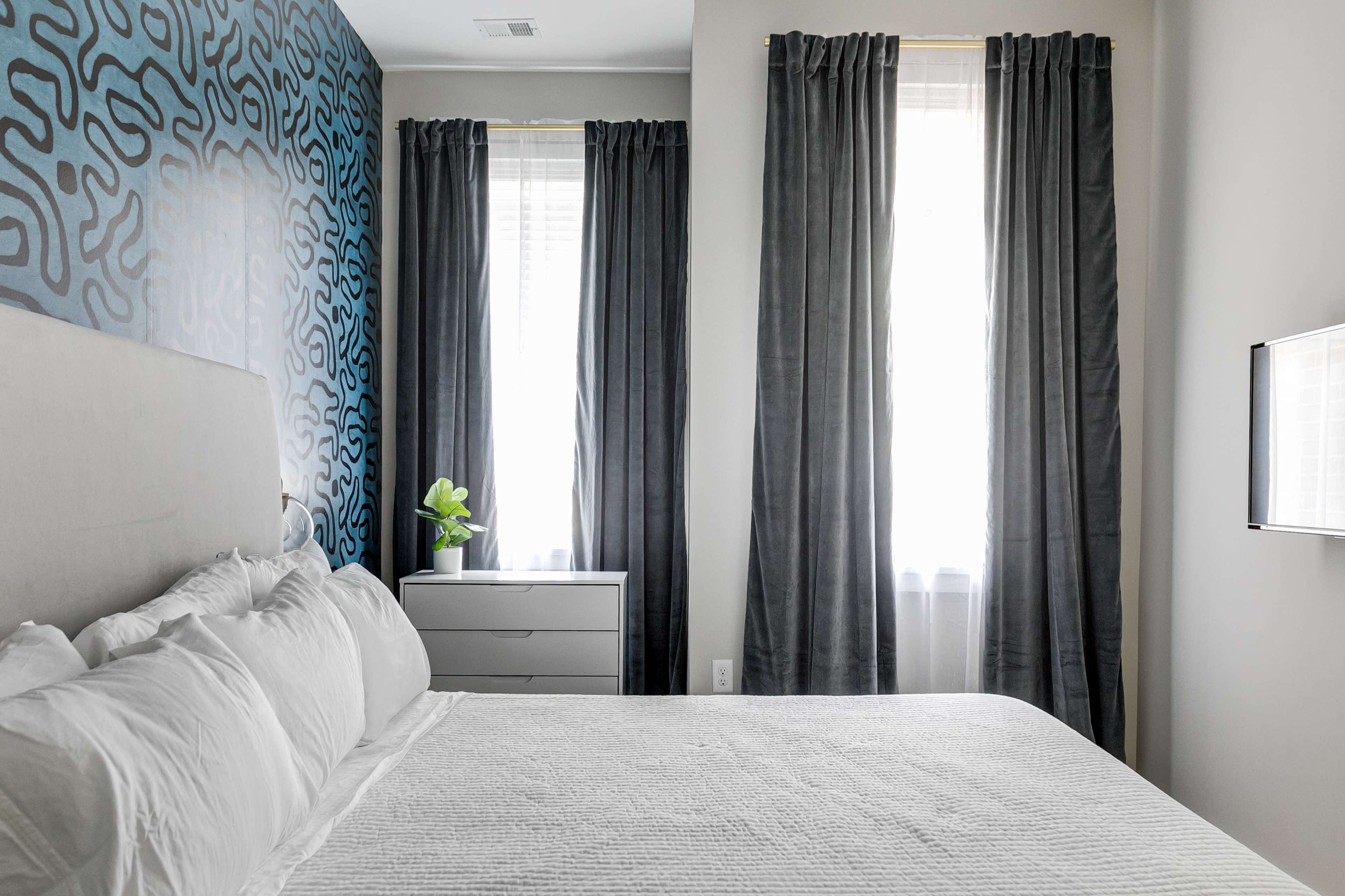 A neatly arranged bedroom with a bed, a modern dresser, and large windows with dark curtains.