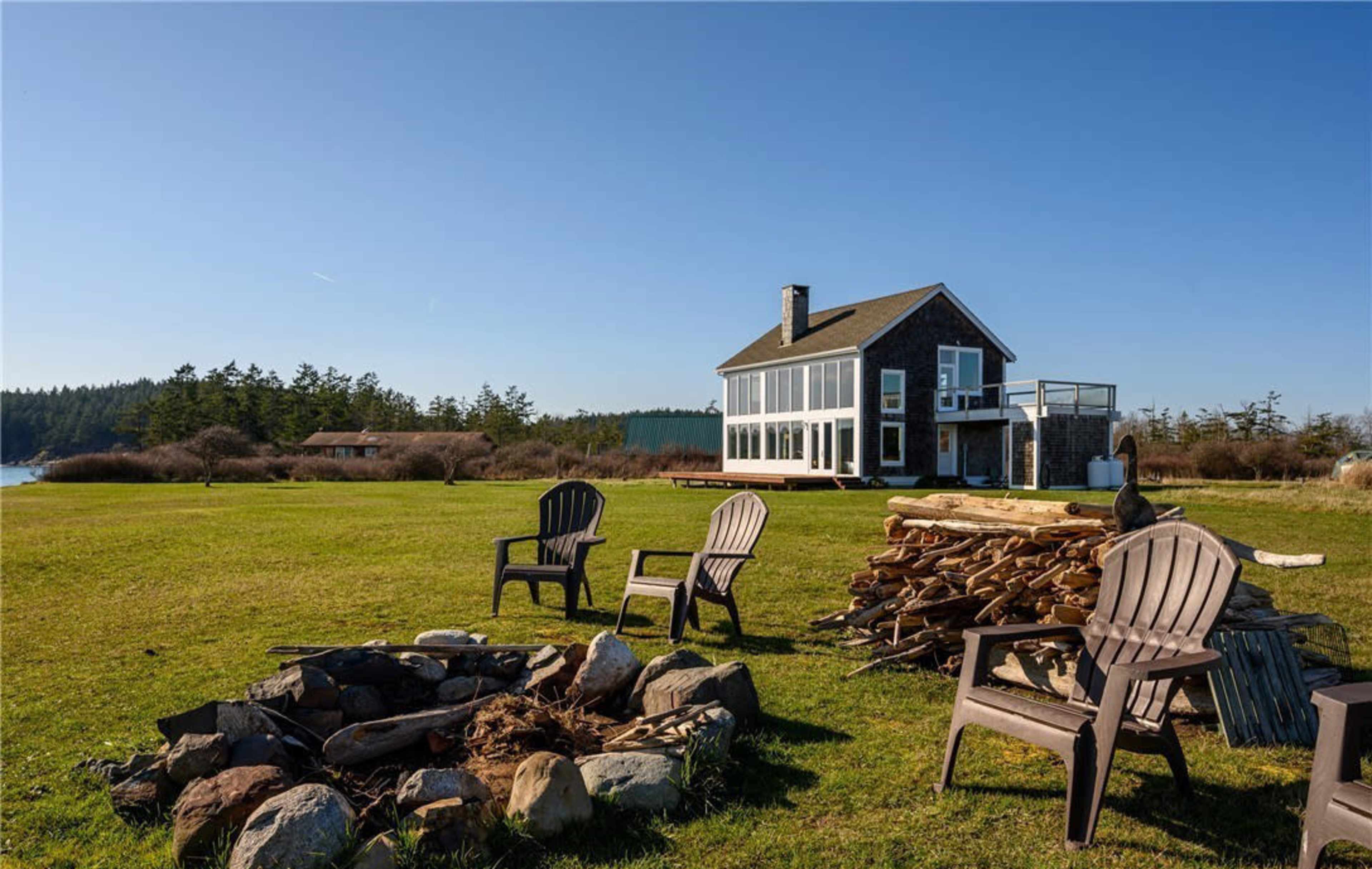 A modern house with large windows overlooks a grassy area containing a fire pit and stacked firewood, set against a clear blue sky and distant trees.