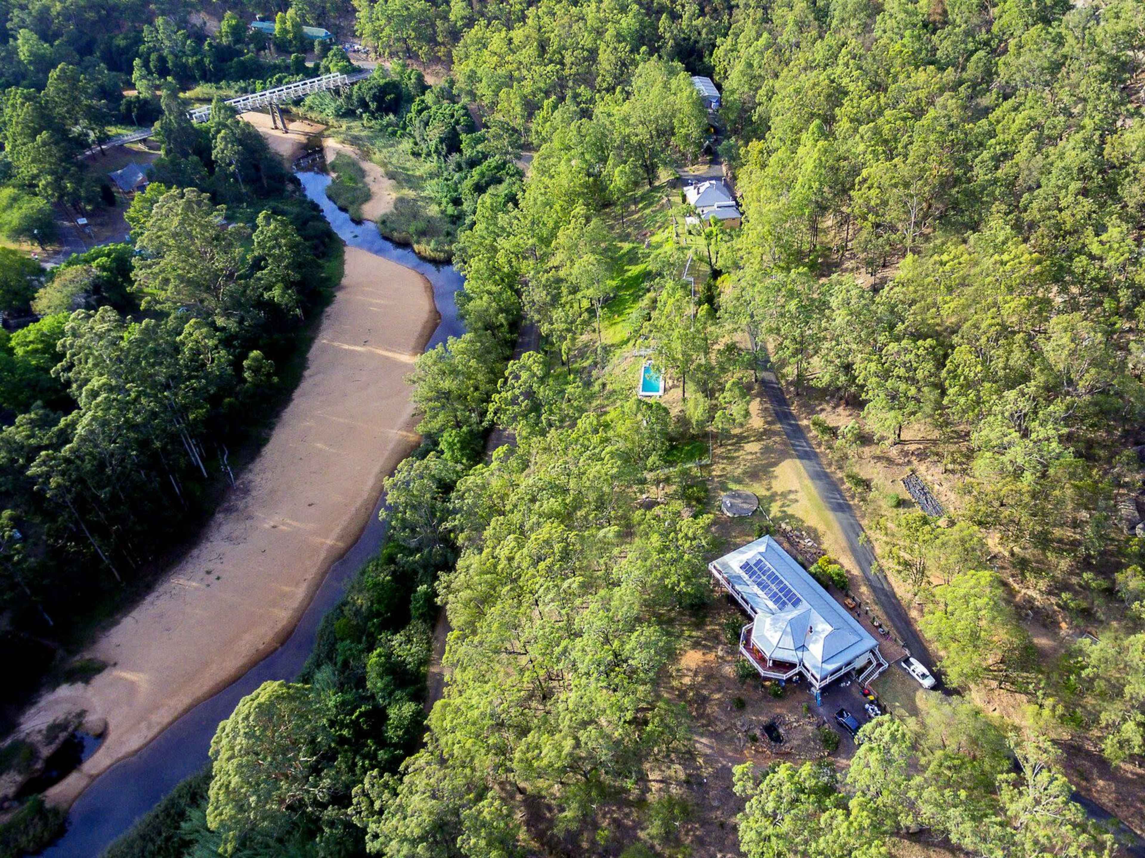 An aerial view of a house surrounded by dense greenery, with a river meandering nearby and a small bridge in the background.