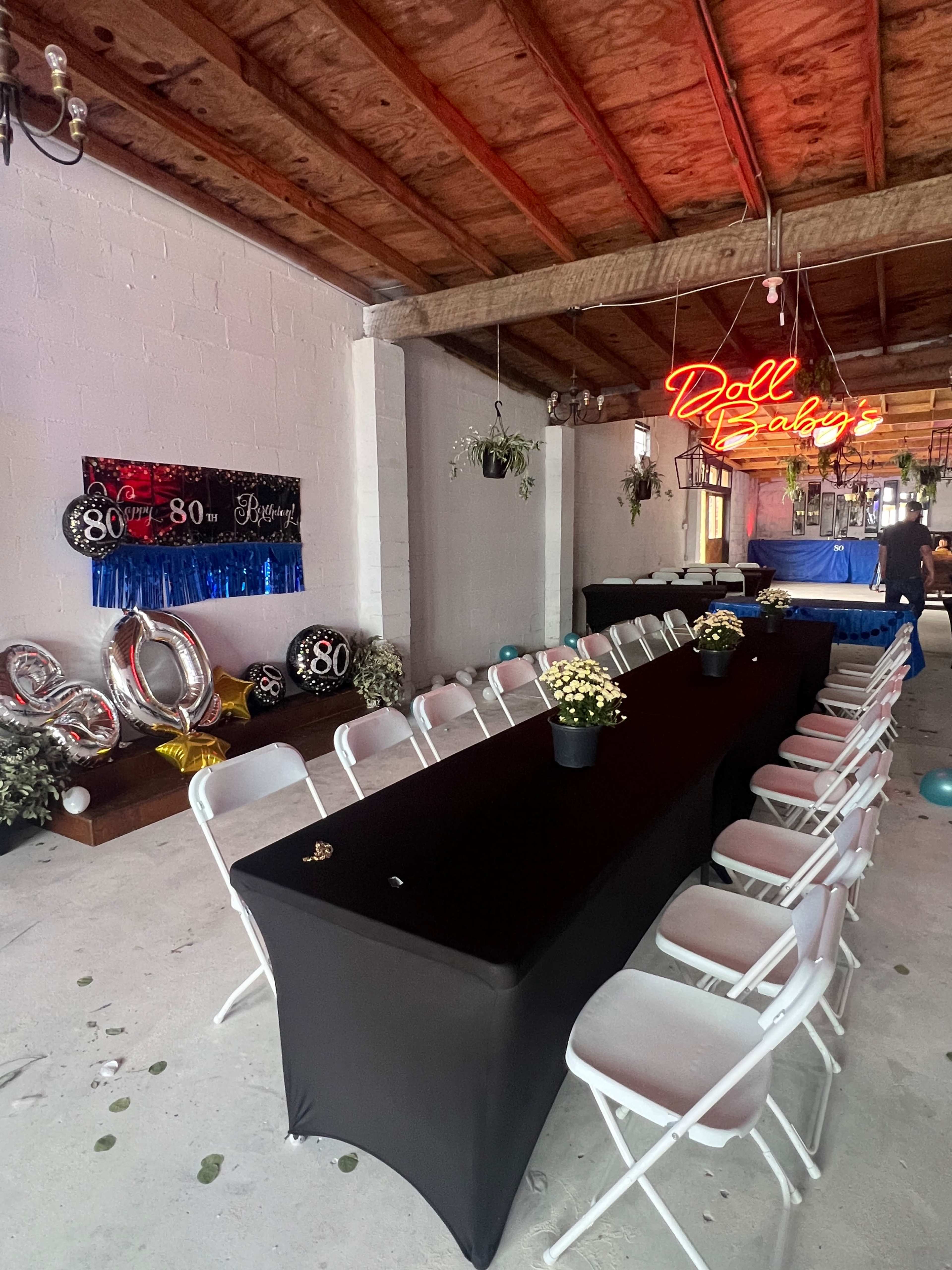 The image shows a decorated indoor venue with a long black table set for a party, featuring balloons and a neon sign that reads "Doll Baby’s 80th."