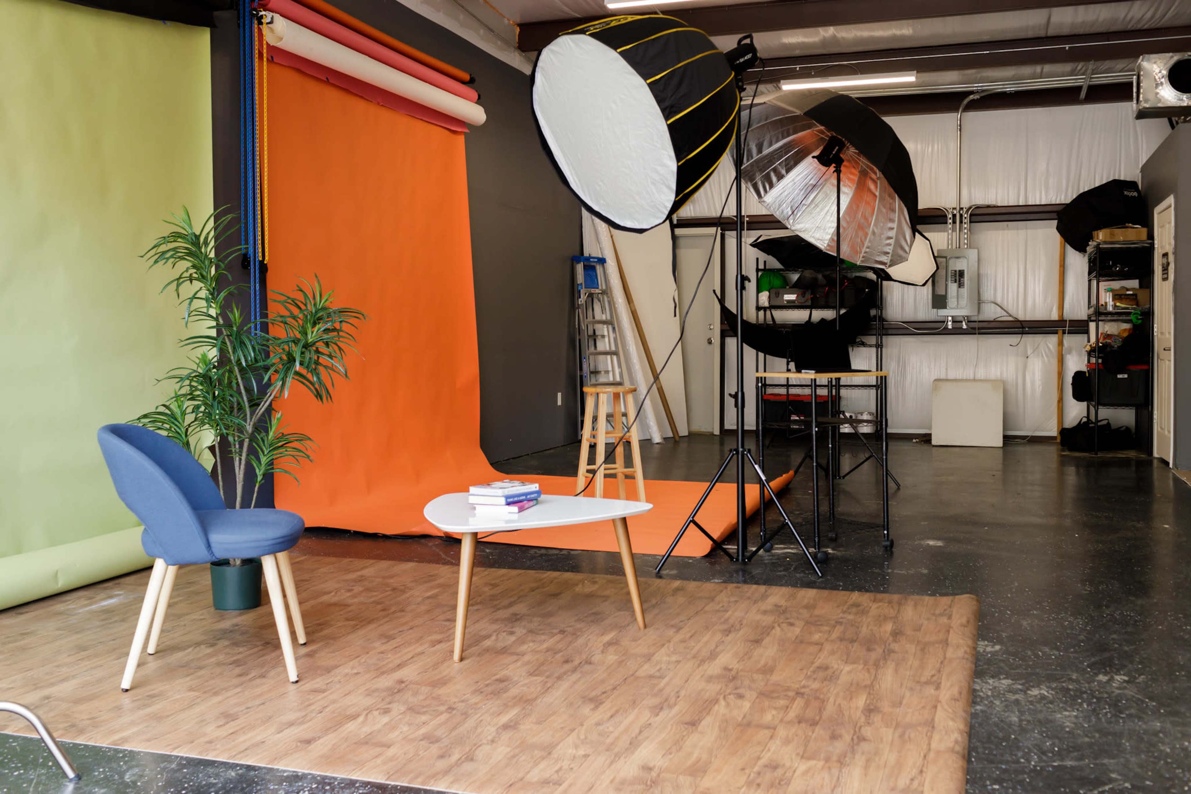 A photography studio with a green and orange backdrop, professional lighting equipment, a blue chair, and a white coffee table with books.