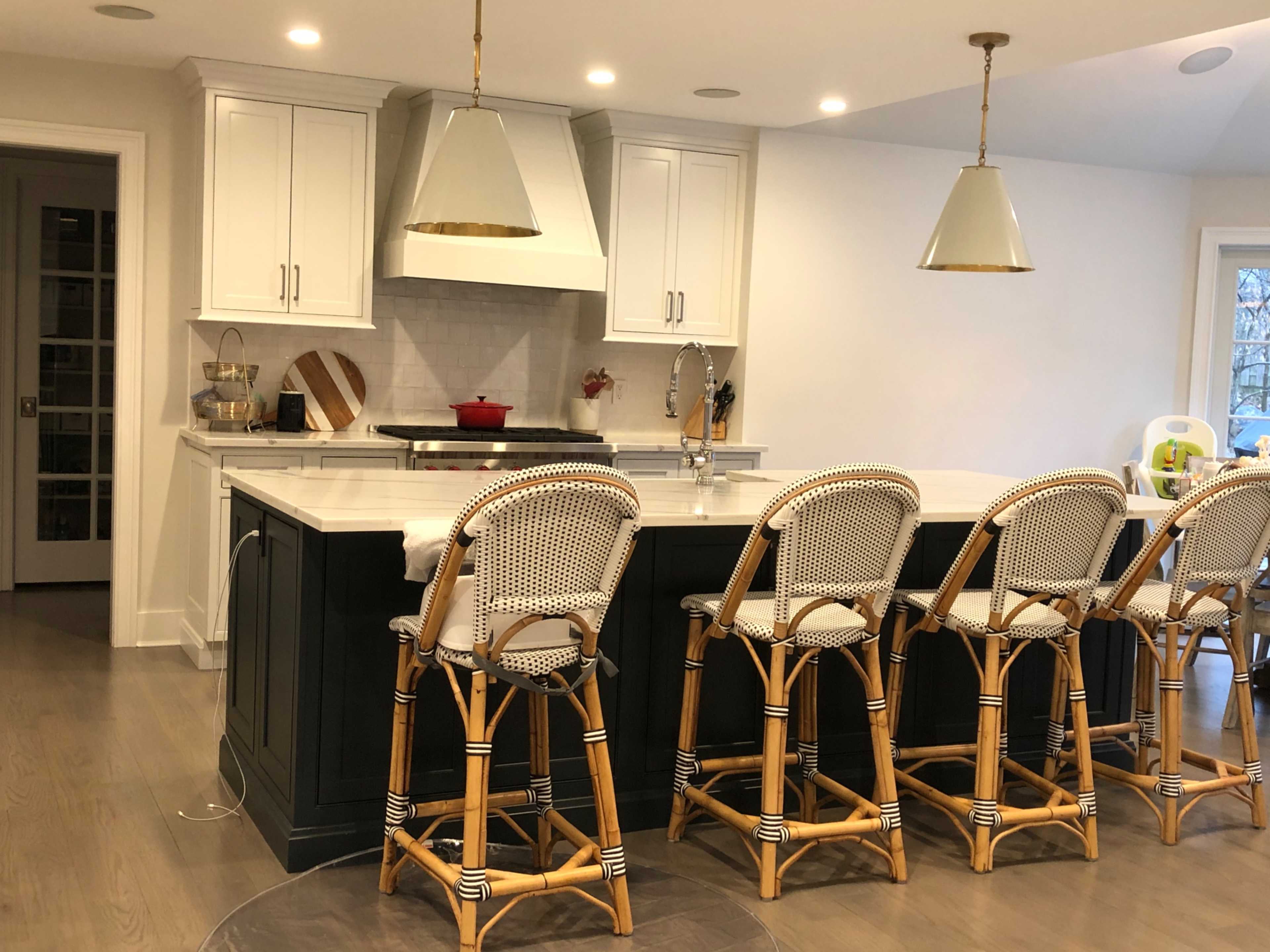 A modern kitchen features a large island with rattan bar stools, white cabinetry, and pendant lighting above.